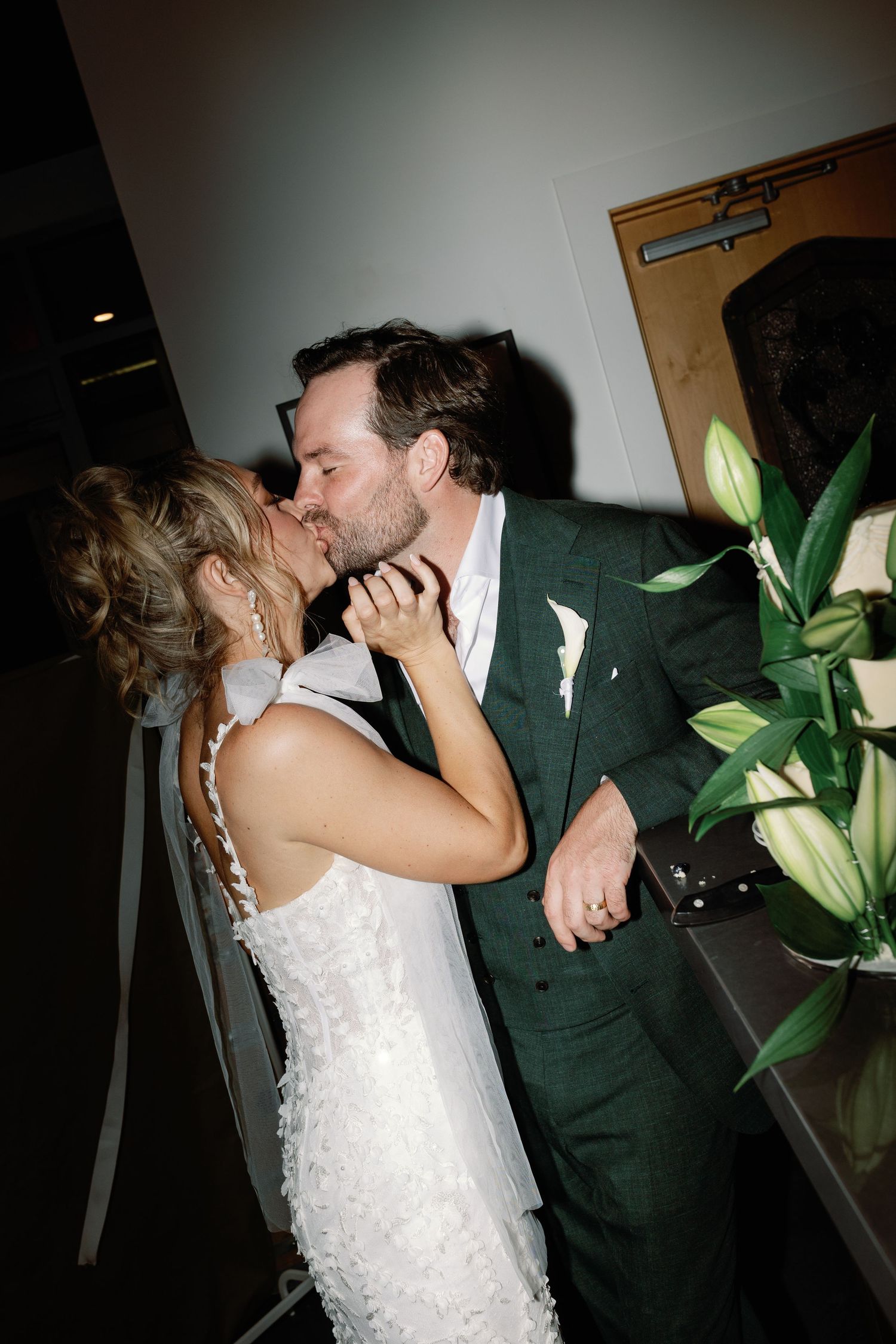 Bride and groom share a romantic kiss near floral arrangement at their wedding reception.