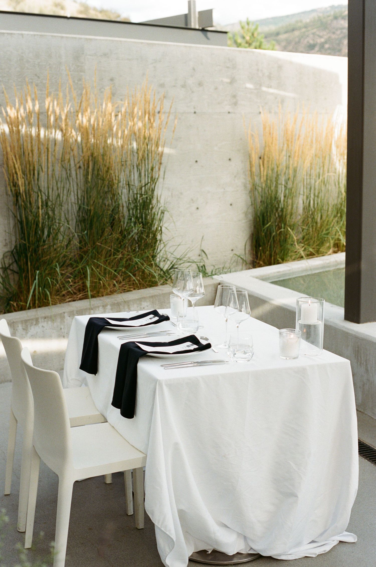 Elegant minimalist wedding reception table set with white linen, black napkins, and glassware near ornamental grasses.
