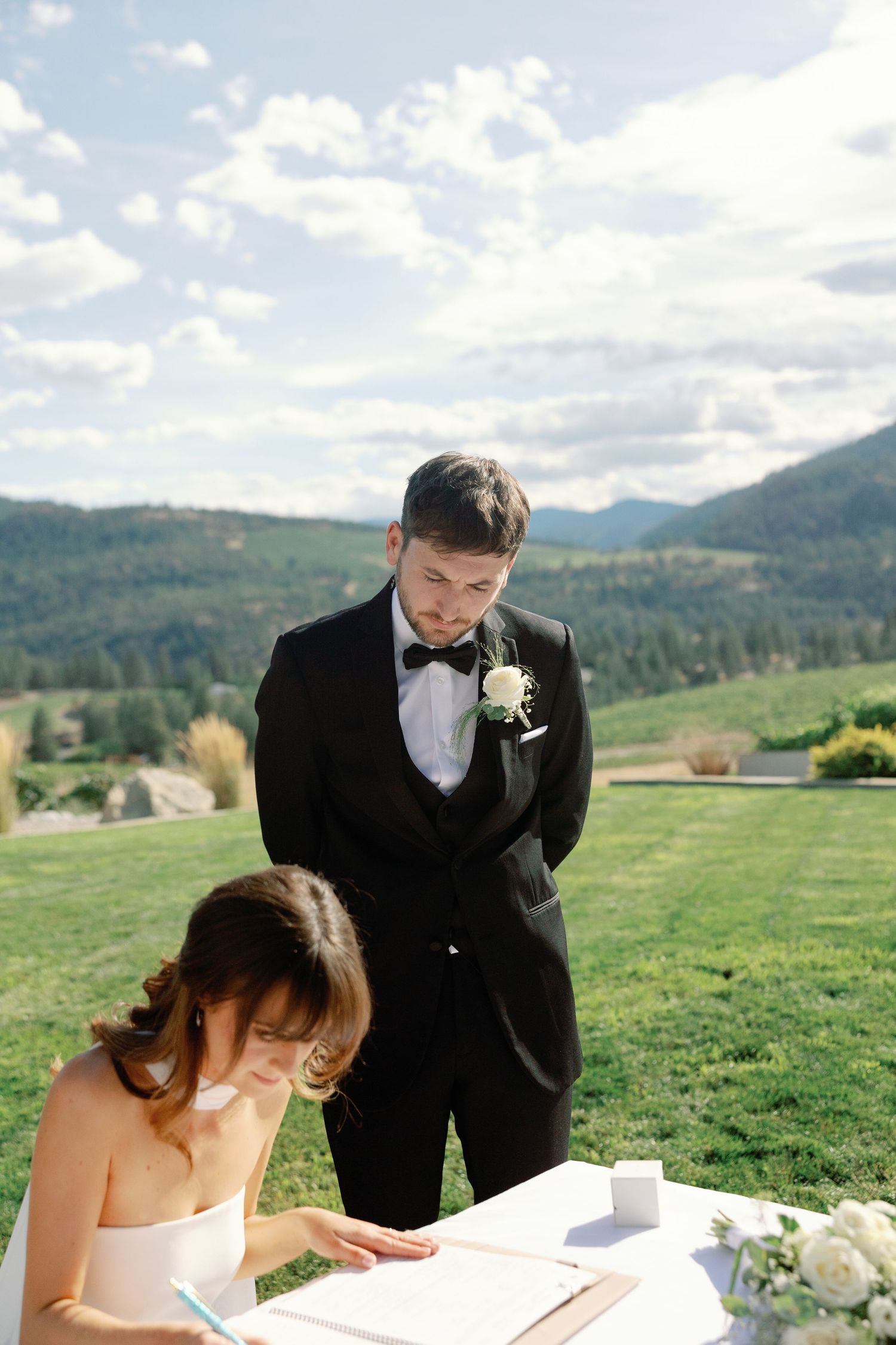 Bride signing wedding registry at outdoor ceremony table while groom in tuxedo watches.