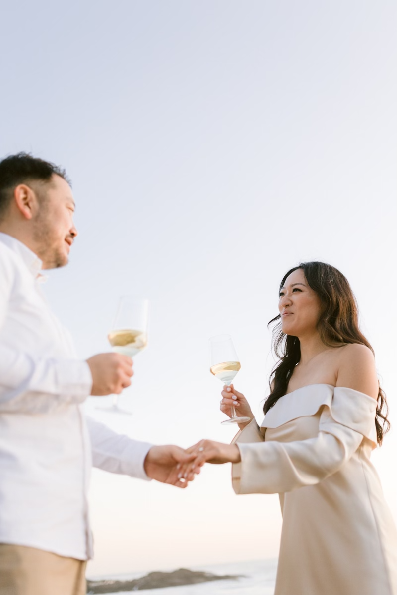 Couple toasting with white wine glasses on a beach at sunset, holding hands in elegant white outfits.