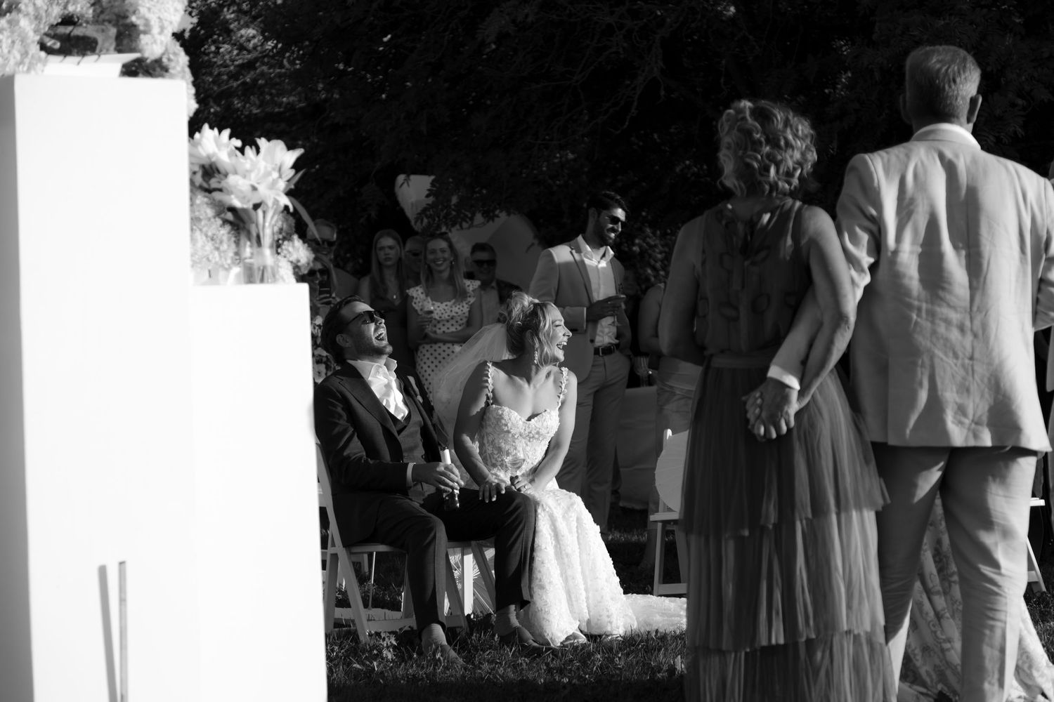 Black and white photo of bride and groom seated during outdoor wedding ceremony.