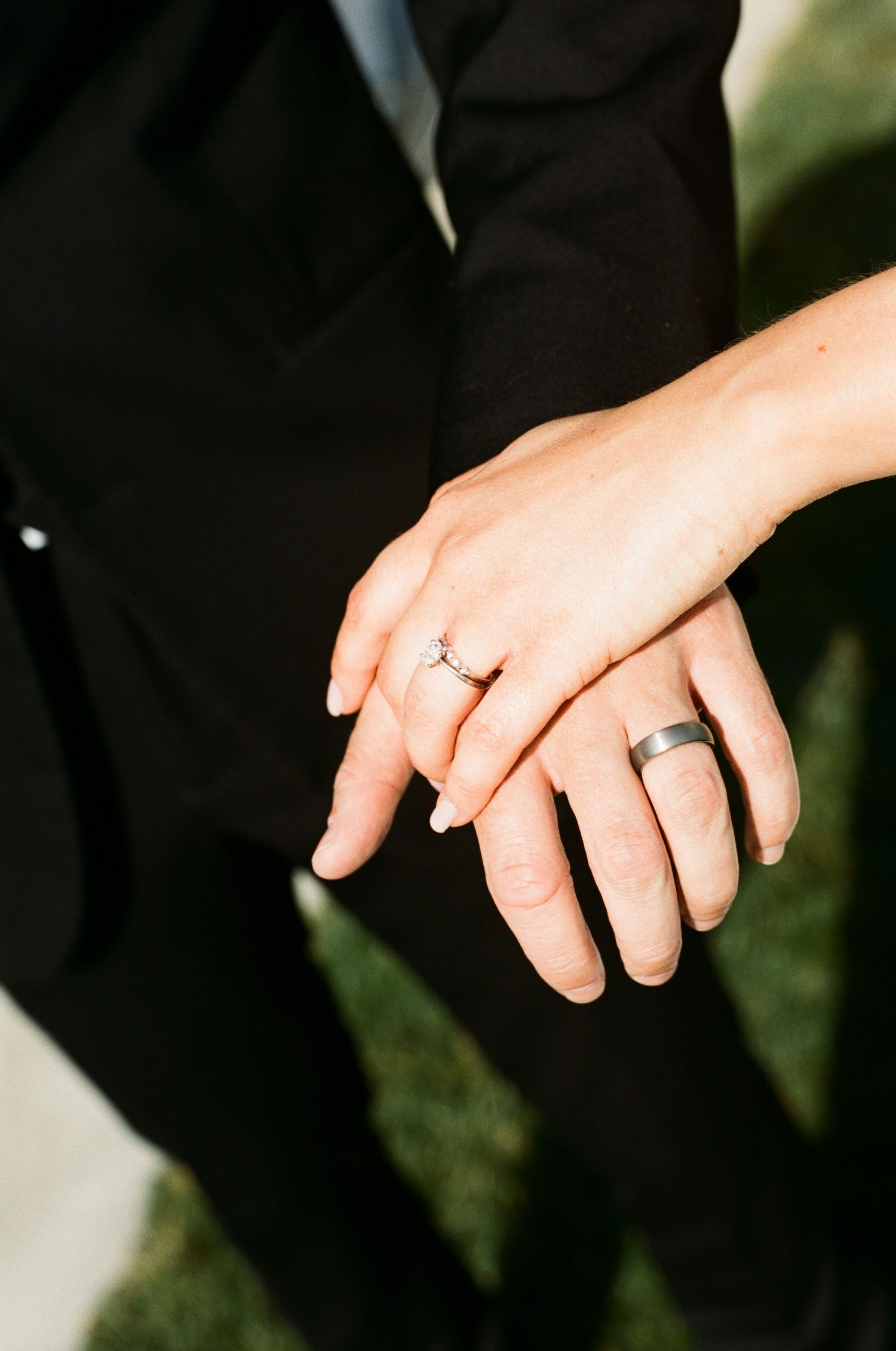 Close-up of newlyweds hands showing diamond engagement ring and silver wedding band against black attire.