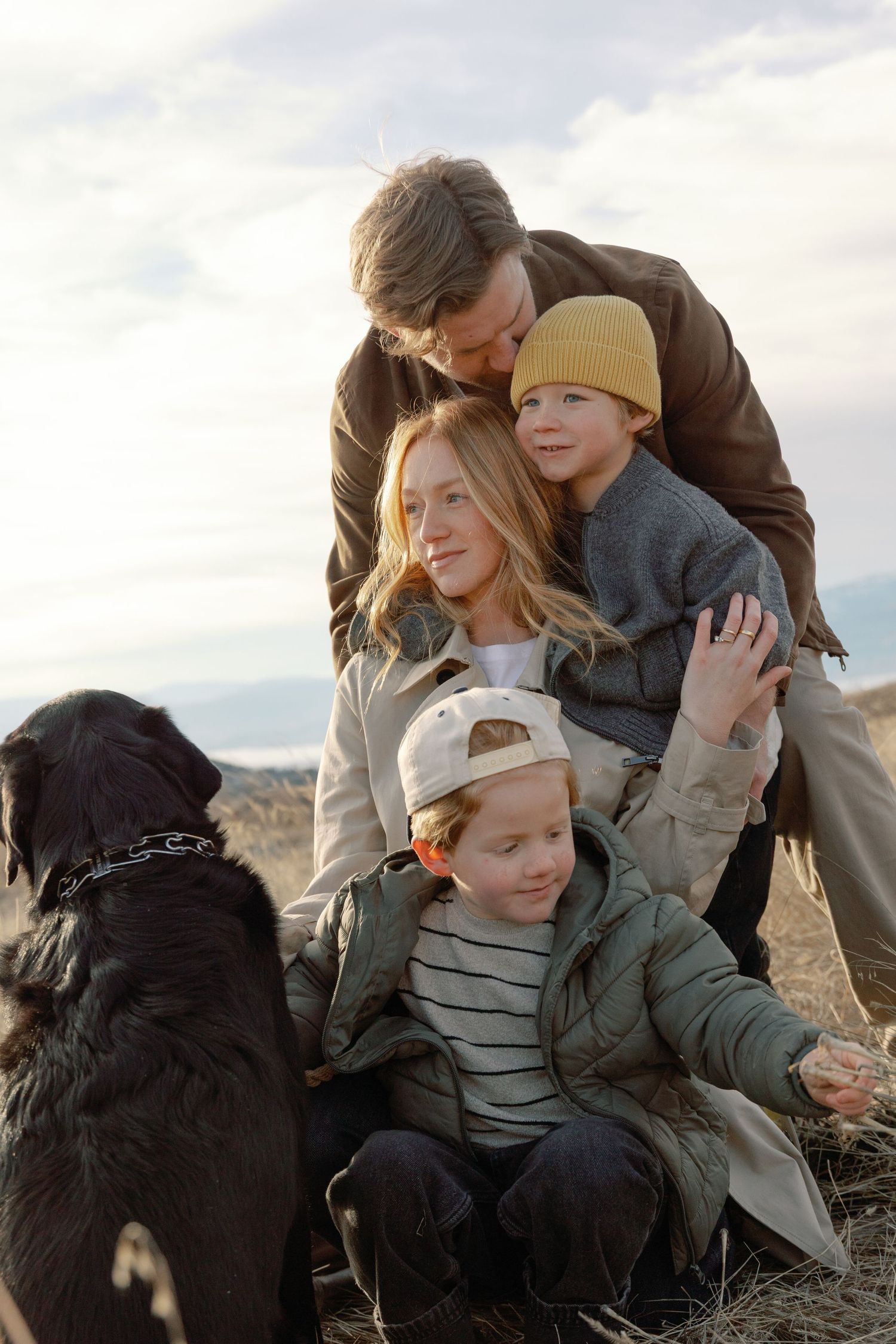 Family of four with black dog posing outdoors in autumn attire, father kissing son's head affectionately.