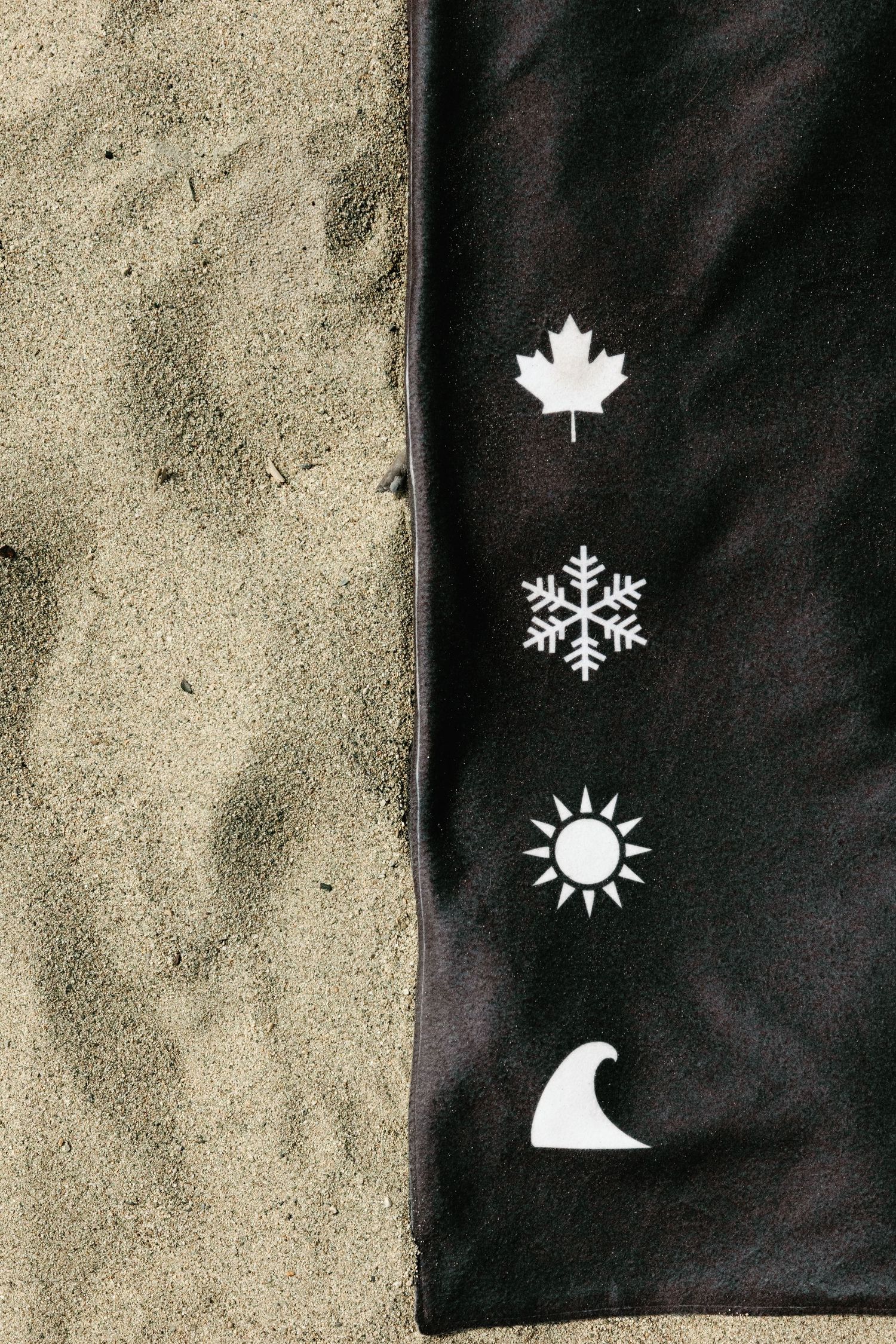 Black fabric on sand with white icons: maple leaf, snowflake, sun, and wave, representing Canadian seasons.