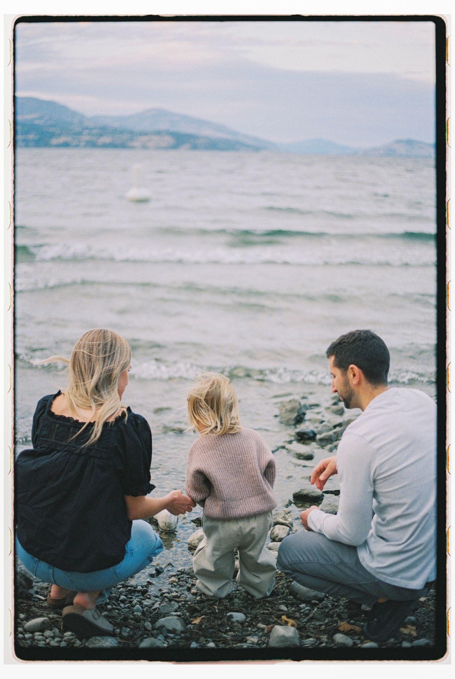 Family of three sitting on rocky beach shore, gazing at ocean waves with mountains in the distance.