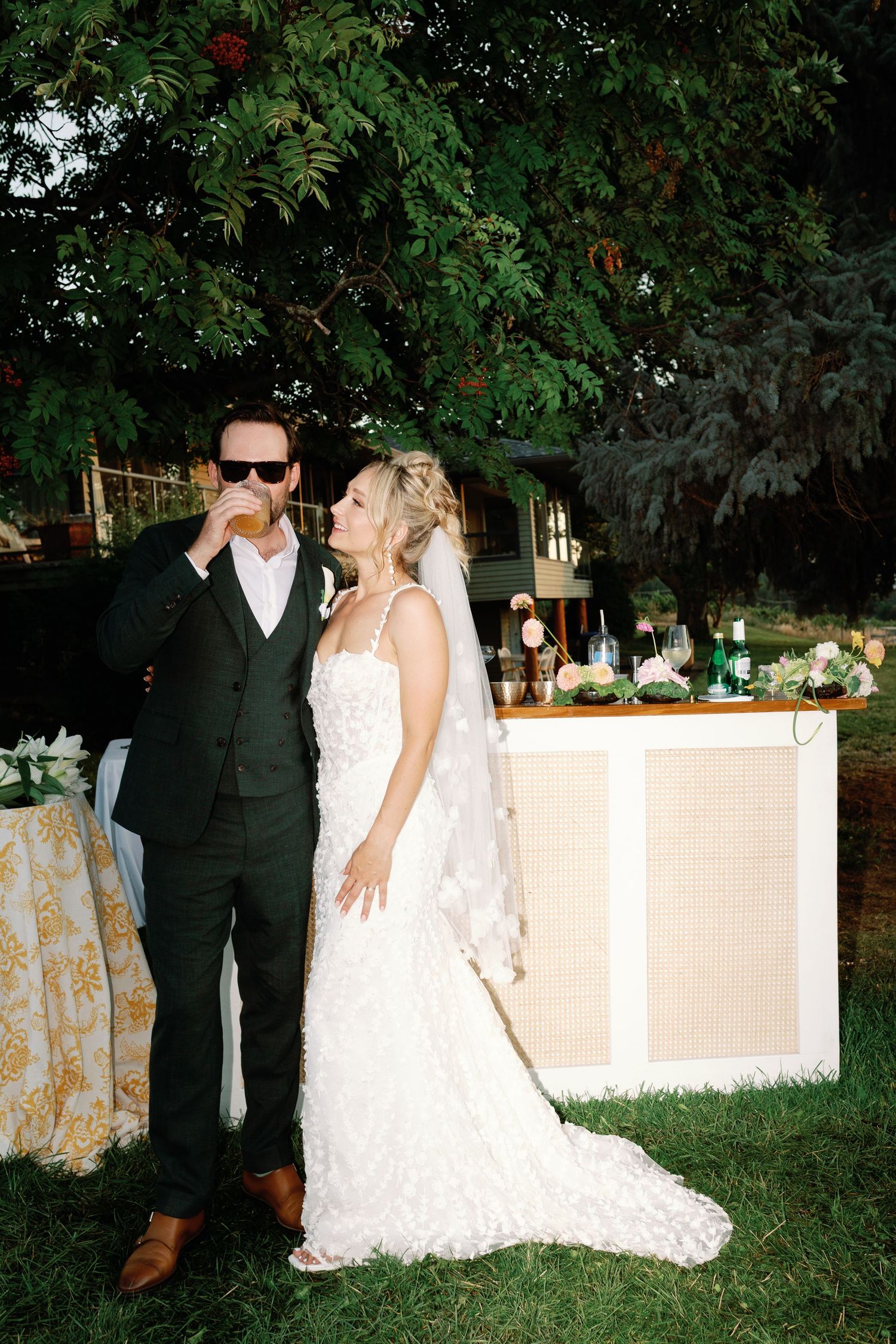 Bride and groom posing by outdoor bar, groom in dark suit with sunglasses, bride in lace wedding gown.