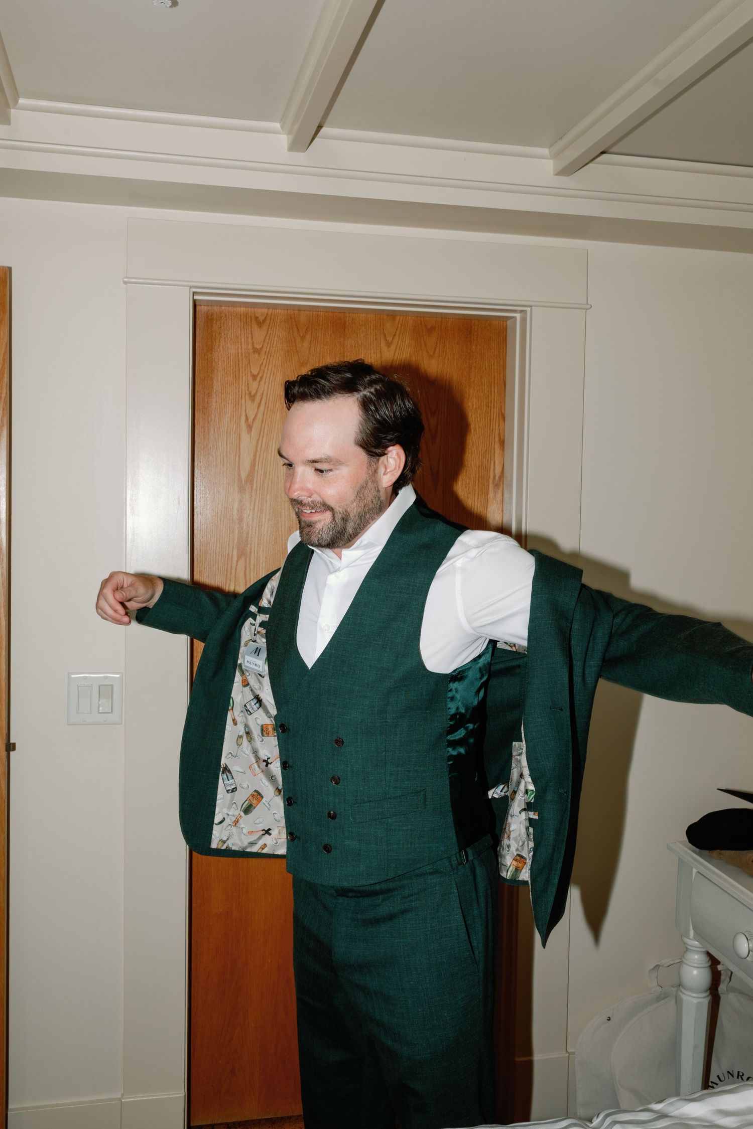 Groom in green vest and patterned suit lining stretches arms while getting dressed in a bright room.