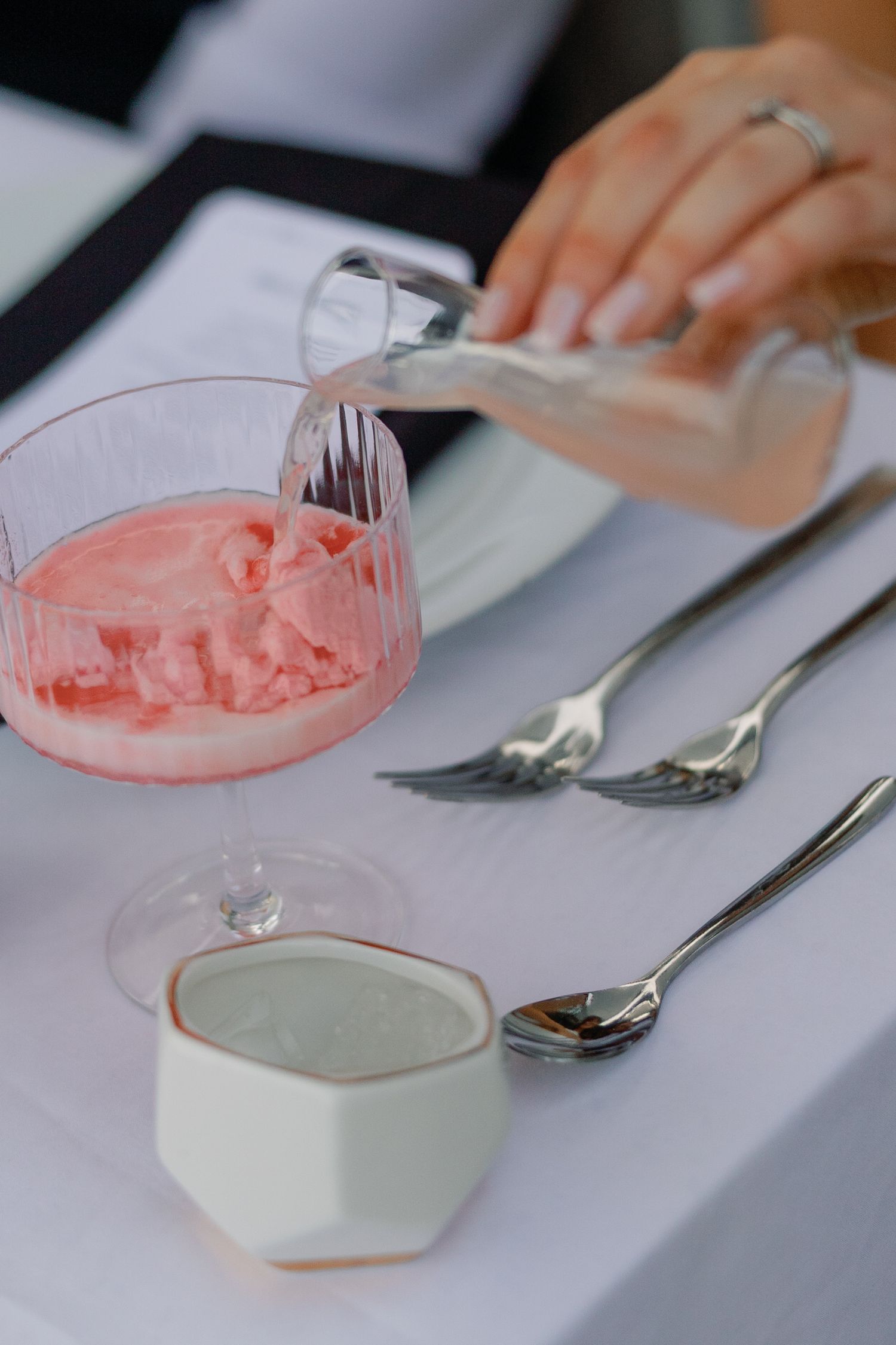 Pink sorbet dessert in crystal glass with silver forks and ceramic creamer on white tablecloth.