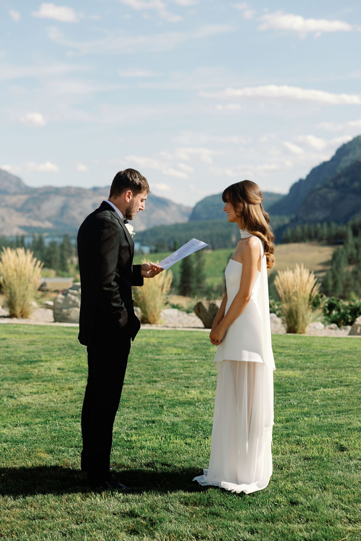 Groom reads wedding vows to bride during outdoor ceremony with mountain backdrop.