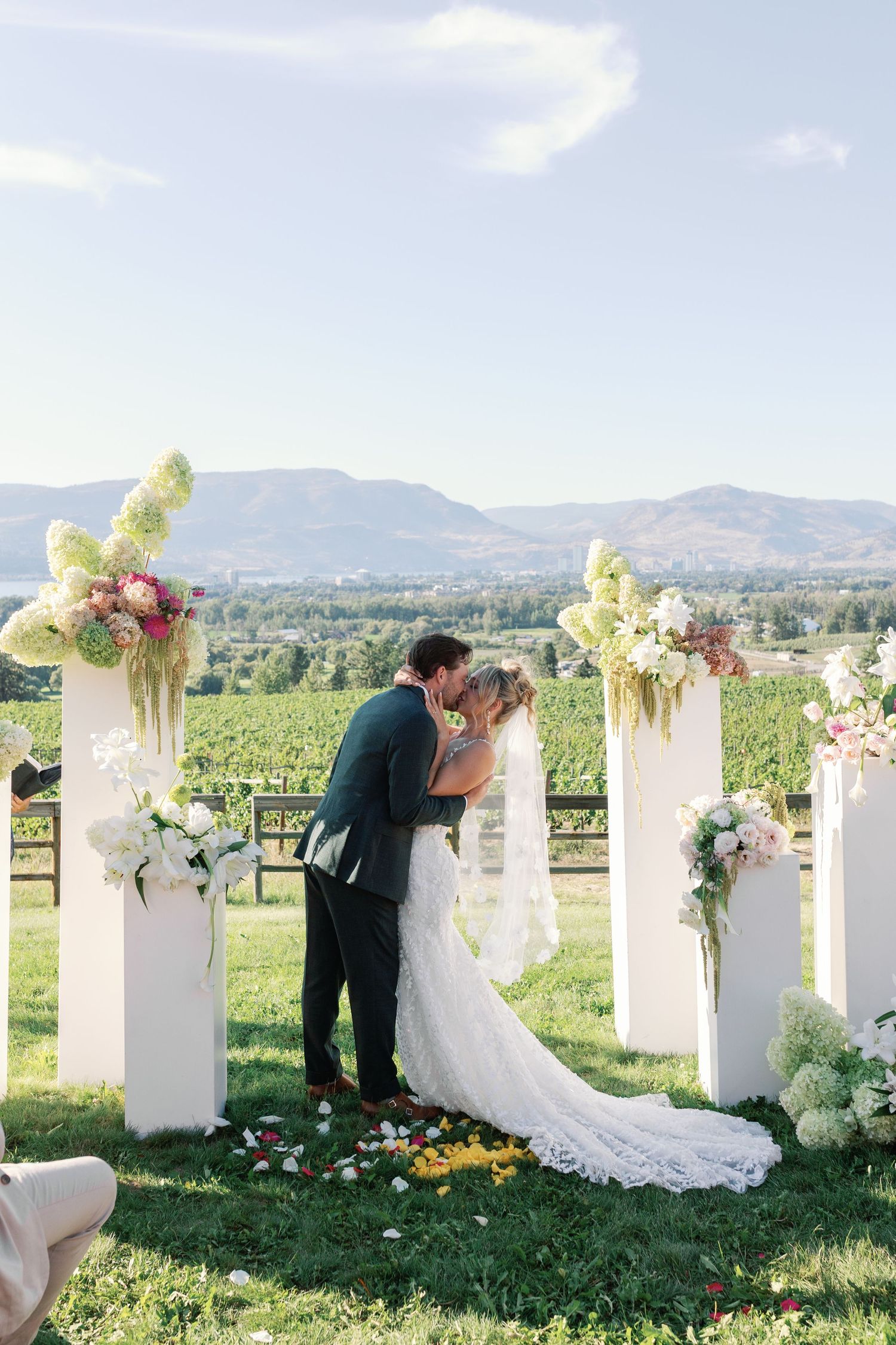 Newlyweds share first kiss at outdoor vineyard wedding altar with mountain backdrop.