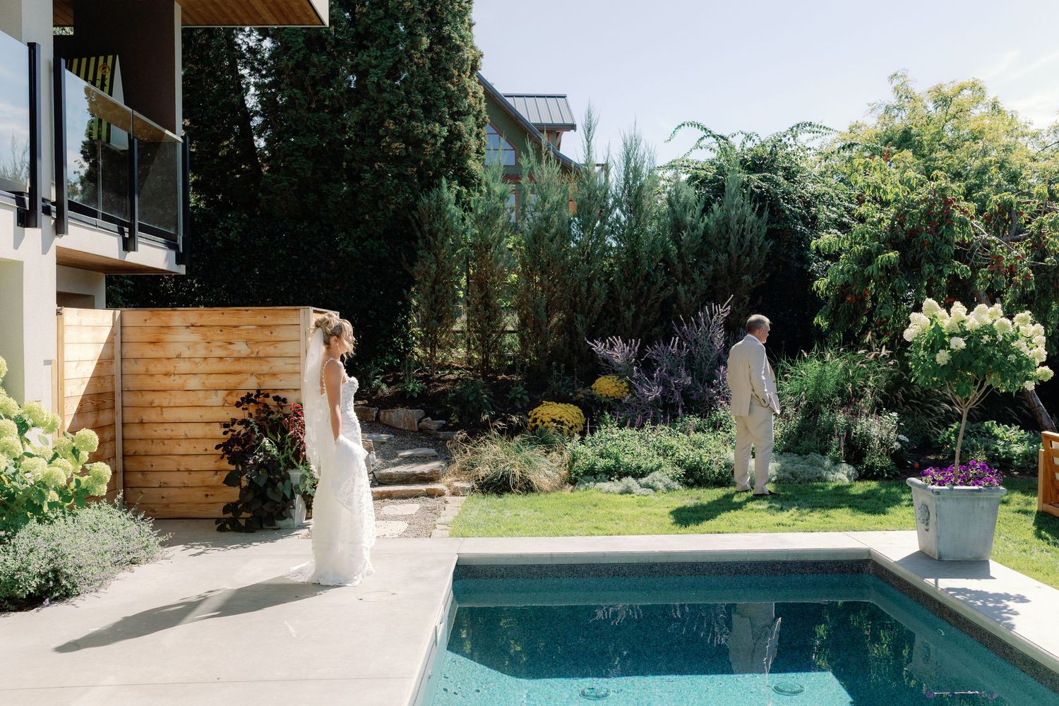Bride in white gown approaches groom for first look moment beside backyard swimming pool surrounded by lush gardens.