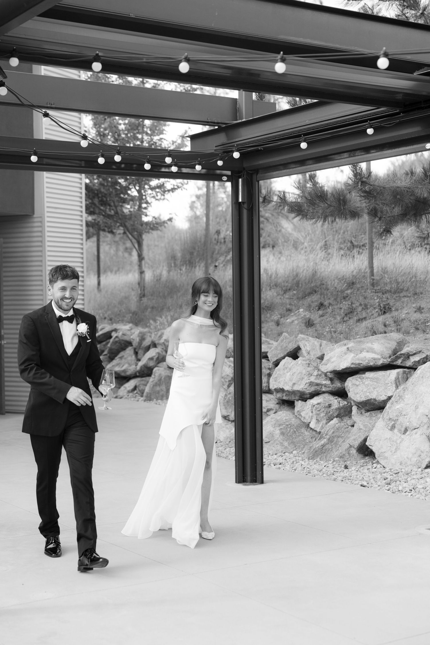 Black and white photo of bride and groom walking together under string lights at elegant outdoor wedding venue.