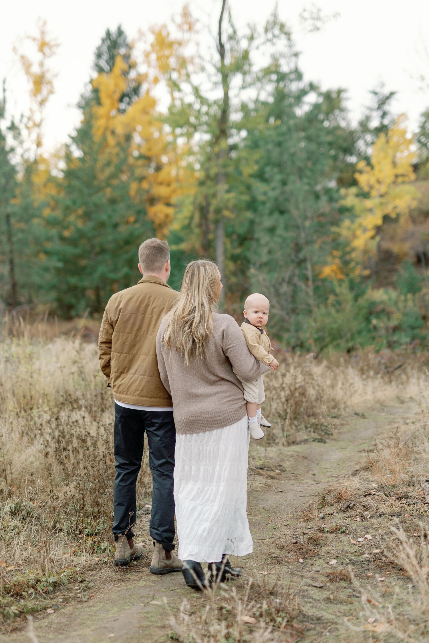 Family of three walking on a dirt path through autumn forest, mother holding baby, father beside her, fall foliage in background.