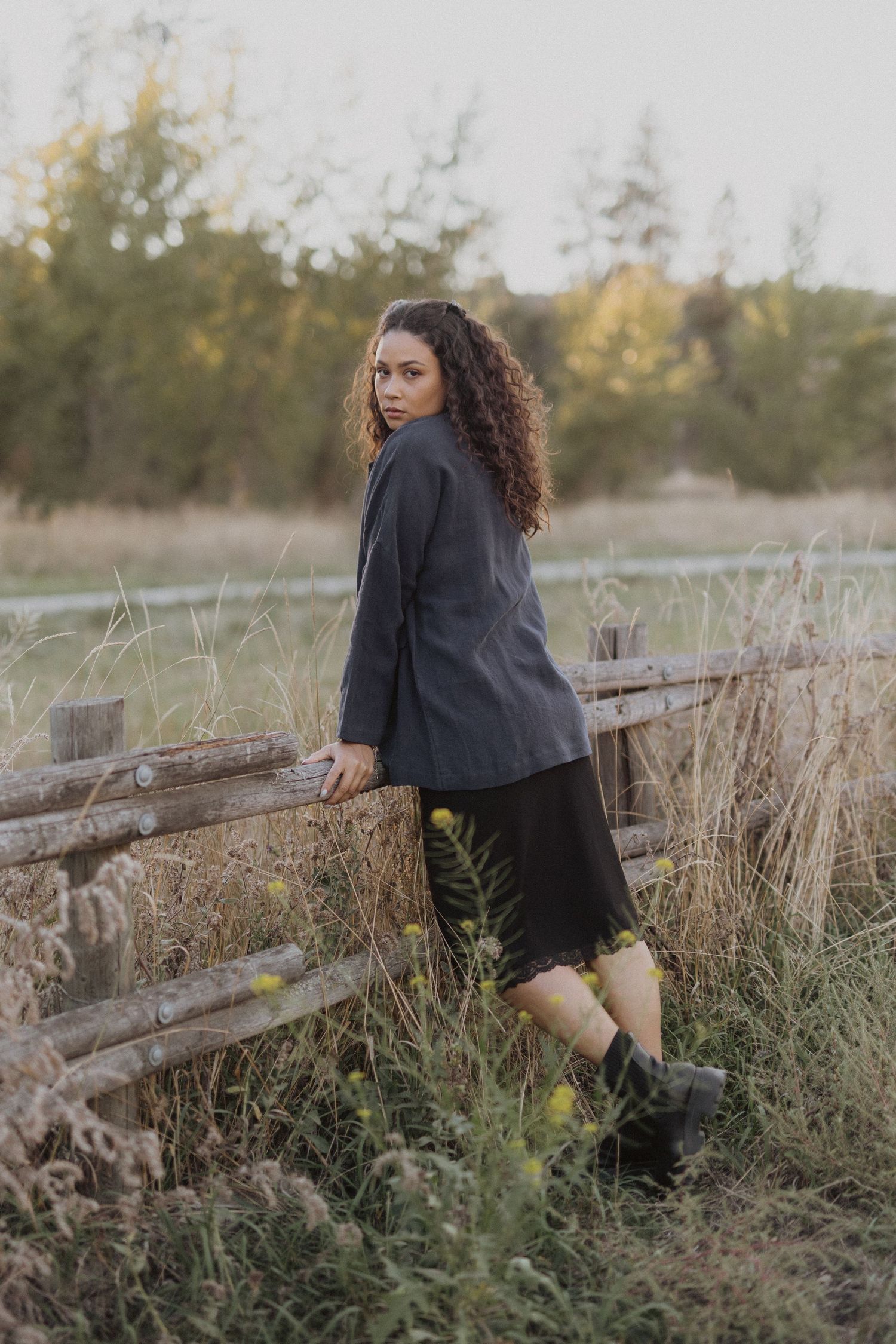 Woman in black blazer and skirt leans on rustic wooden fence in golden field at sunset.