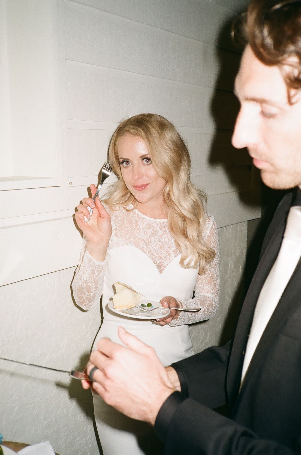 Blonde bride in lace wedding dress eating cake and smiling at reception, film photography style.