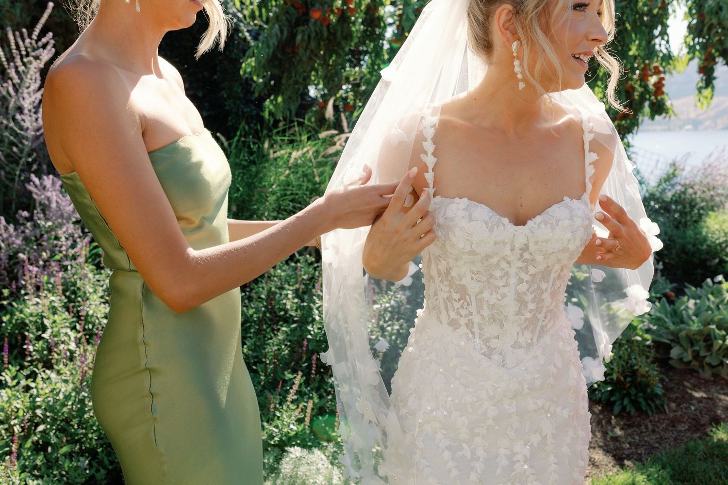Bridesmaid in sage green dress helping bride with lace wedding gown in garden setting.