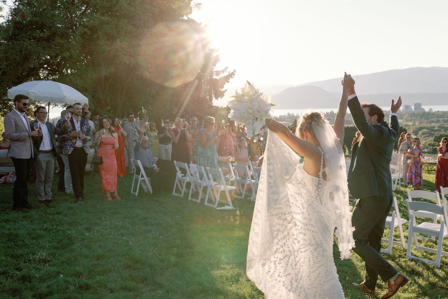 Bride and groom raise hands joyfully as guests cheer during golden hour outdoor wedding ceremony.
