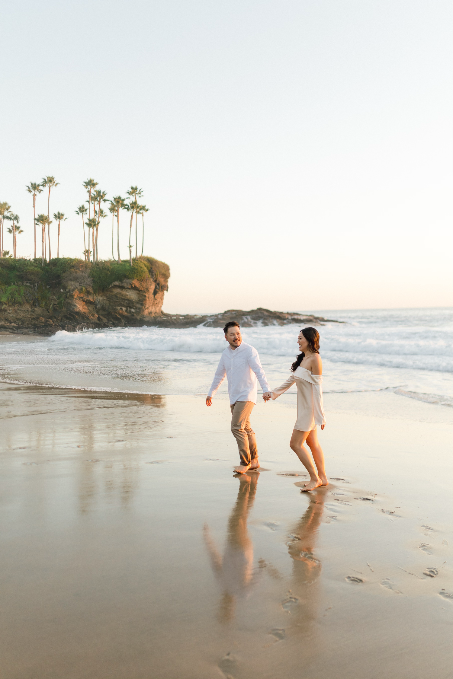 A couple holds hands and walks barefoot along a sunny beach with palm trees and cliffs in the background.