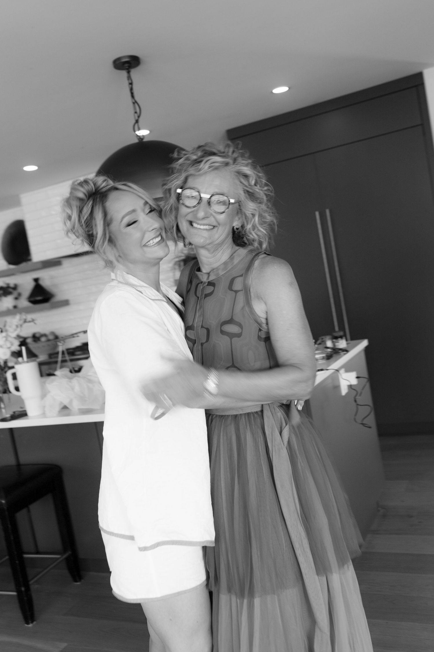 Black and white photo of bride in white pajamas sharing a warm embrace with older woman in patterned dress.