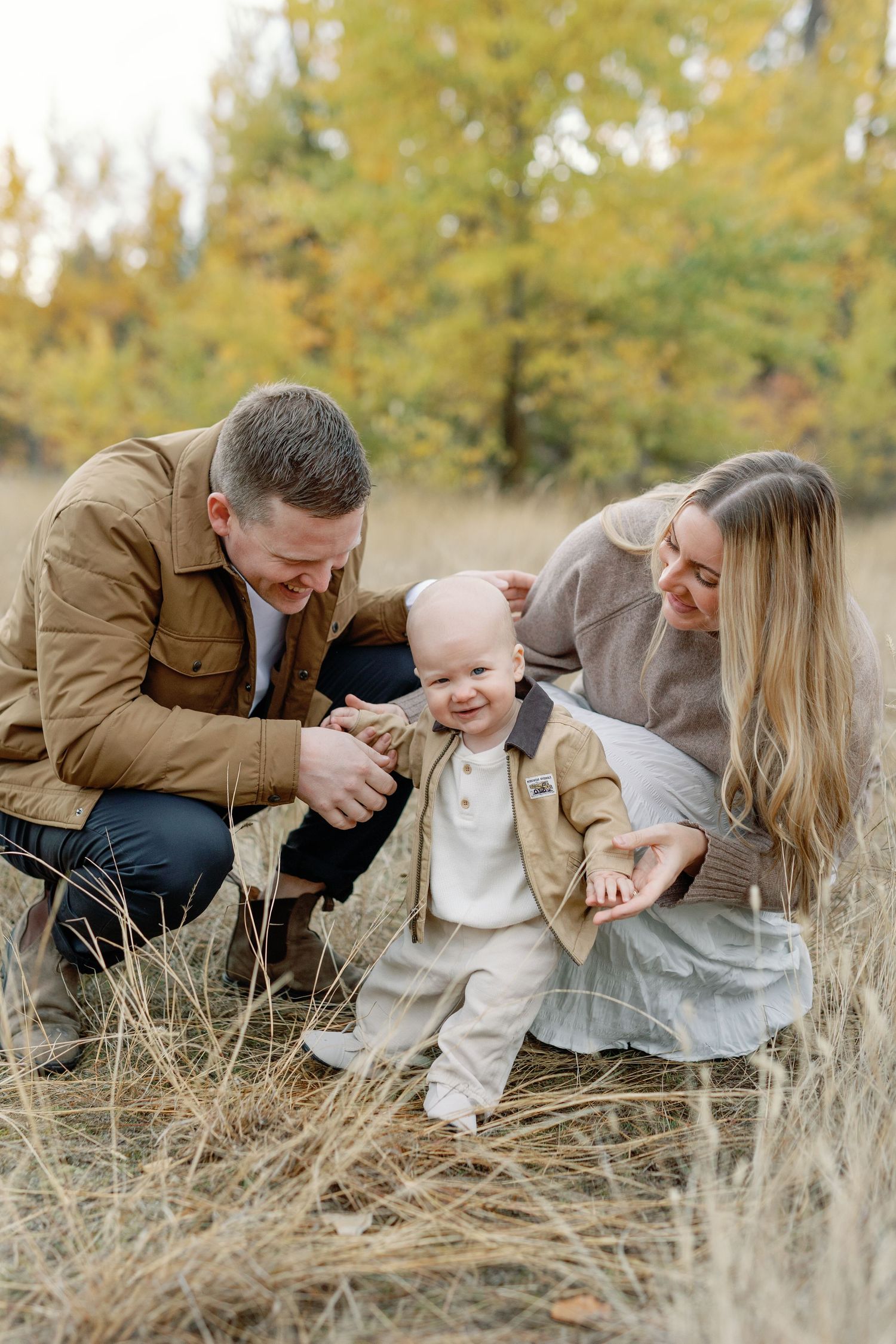 Family of three enjoying autumn outdoors, parents smiling at happy toddler in fall field with colorful trees.