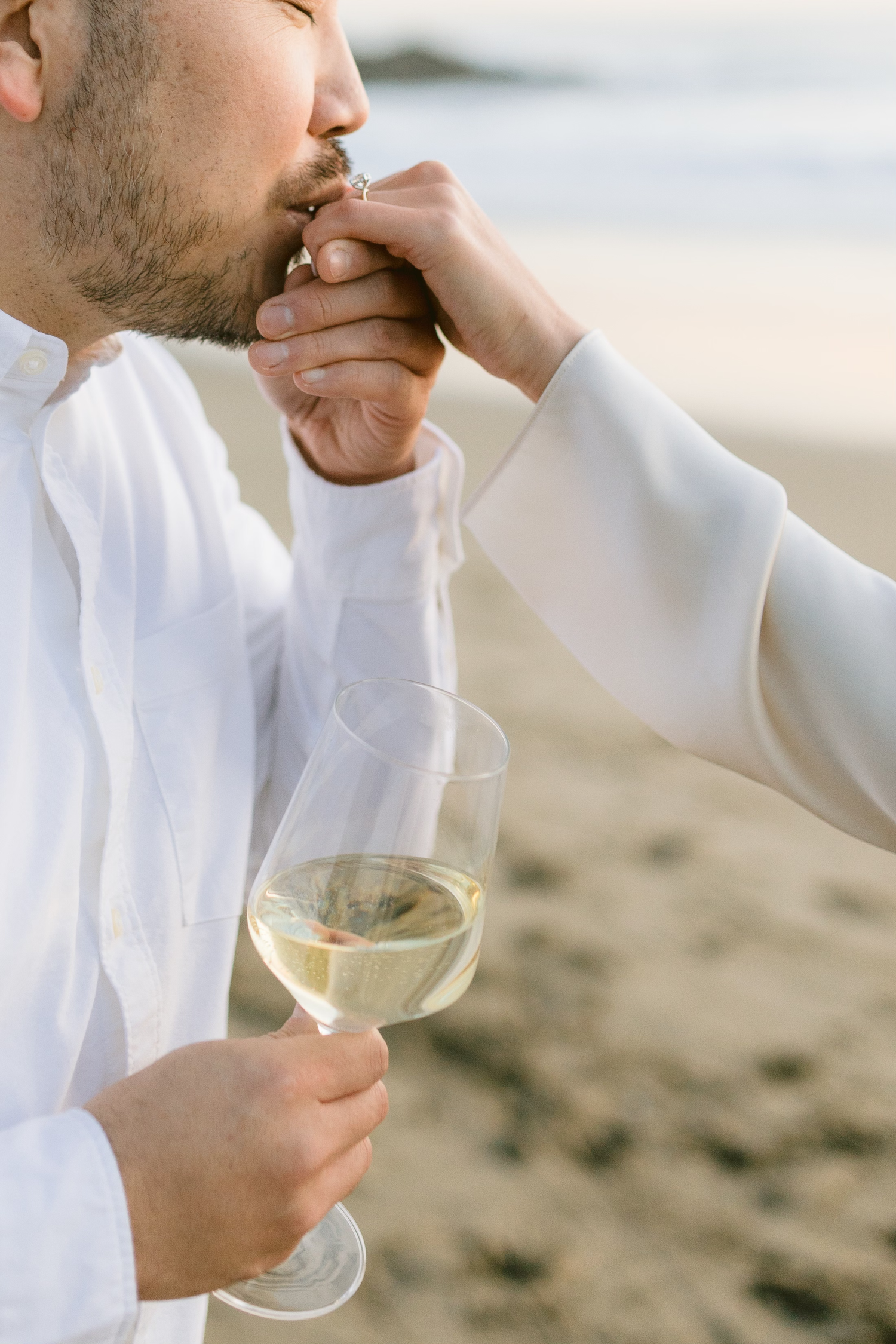 Man in white shirt kissing woman's hand with engagement ring while holding a glass of white wine on beach.