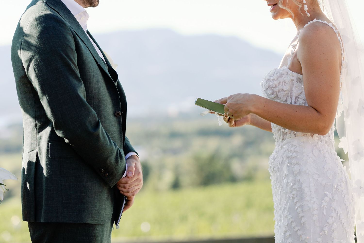 Bride reads wedding vows from card while groom listens at outdoor vineyard ceremony.
