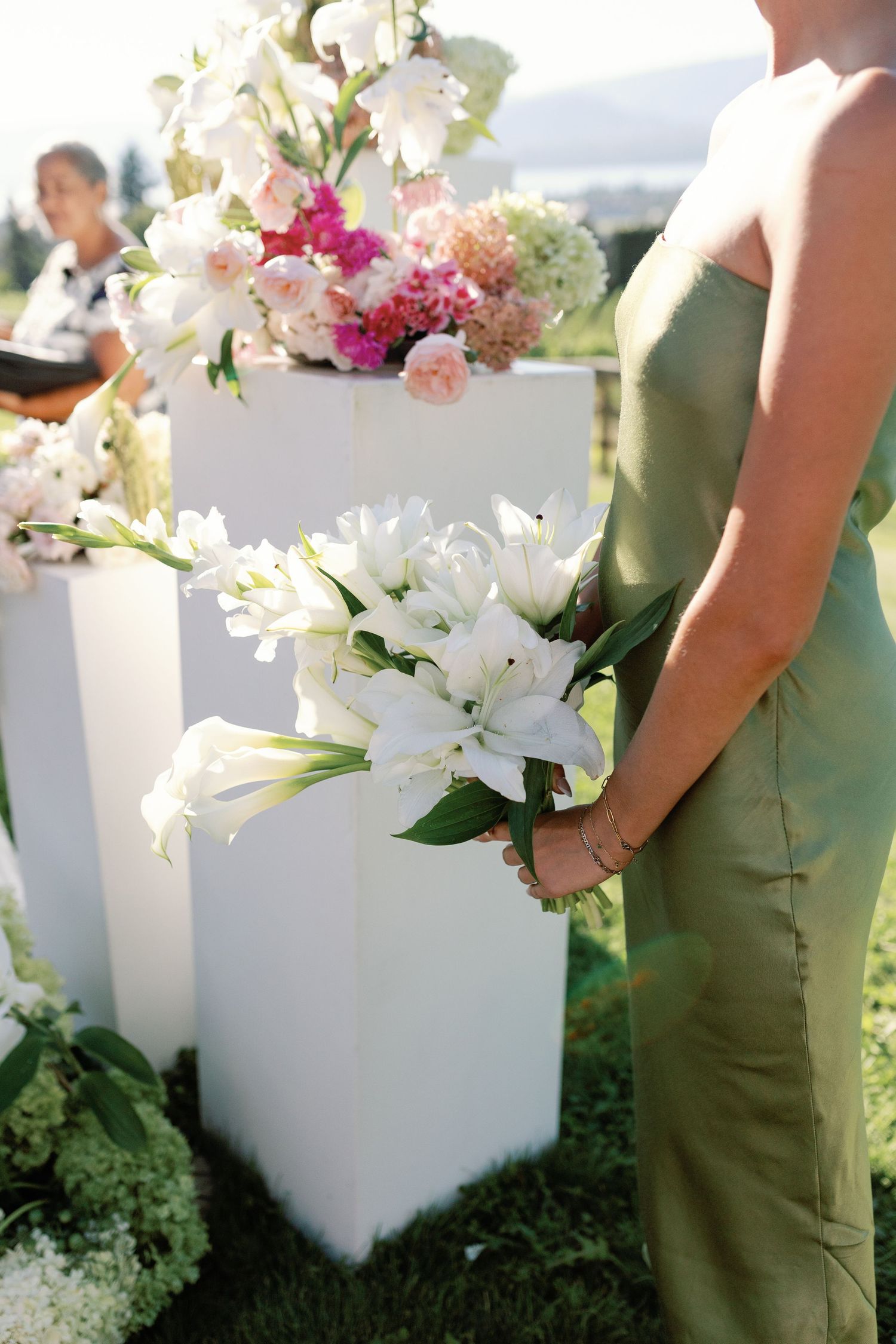 Bridesmaid in green dress holding white lily bouquet at outdoor wedding ceremony with floral pedestals.