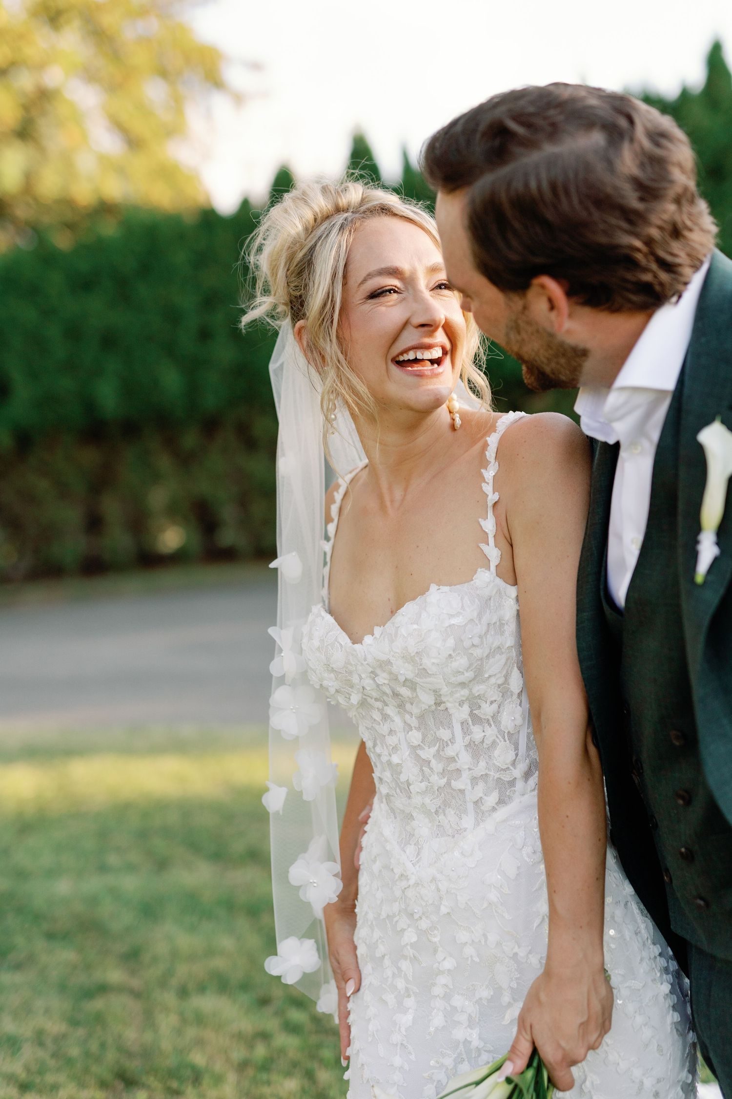 Radiant bride laughs joyfully as groom whispers to her at outdoor wedding.