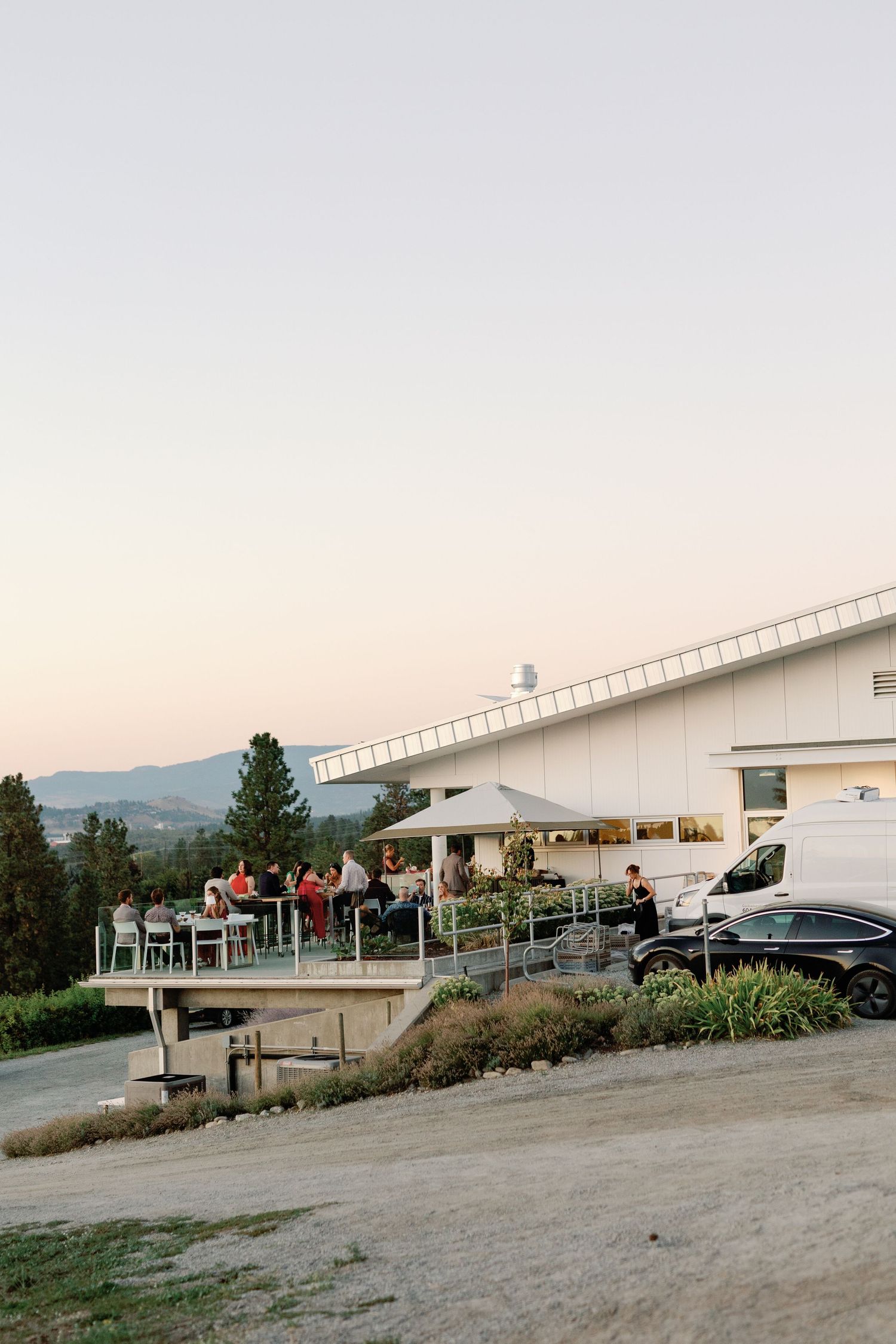 Guests dining on outdoor terrace of modern winery building at sunset with rolling hills in background.