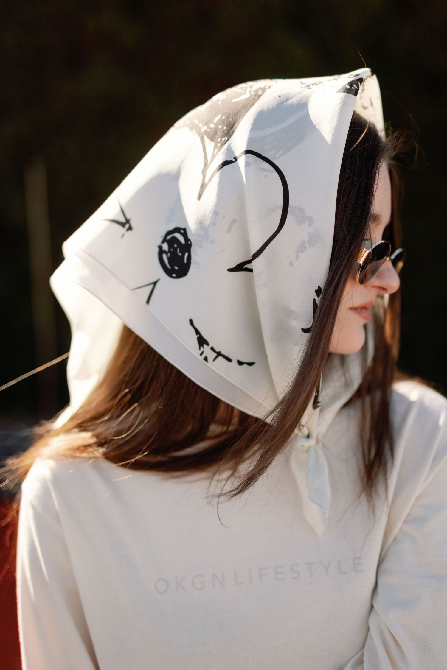Woman wearing white OKGN Lifestyle hoodie with drawn ghost face design on hood, paired with round sunglasses.