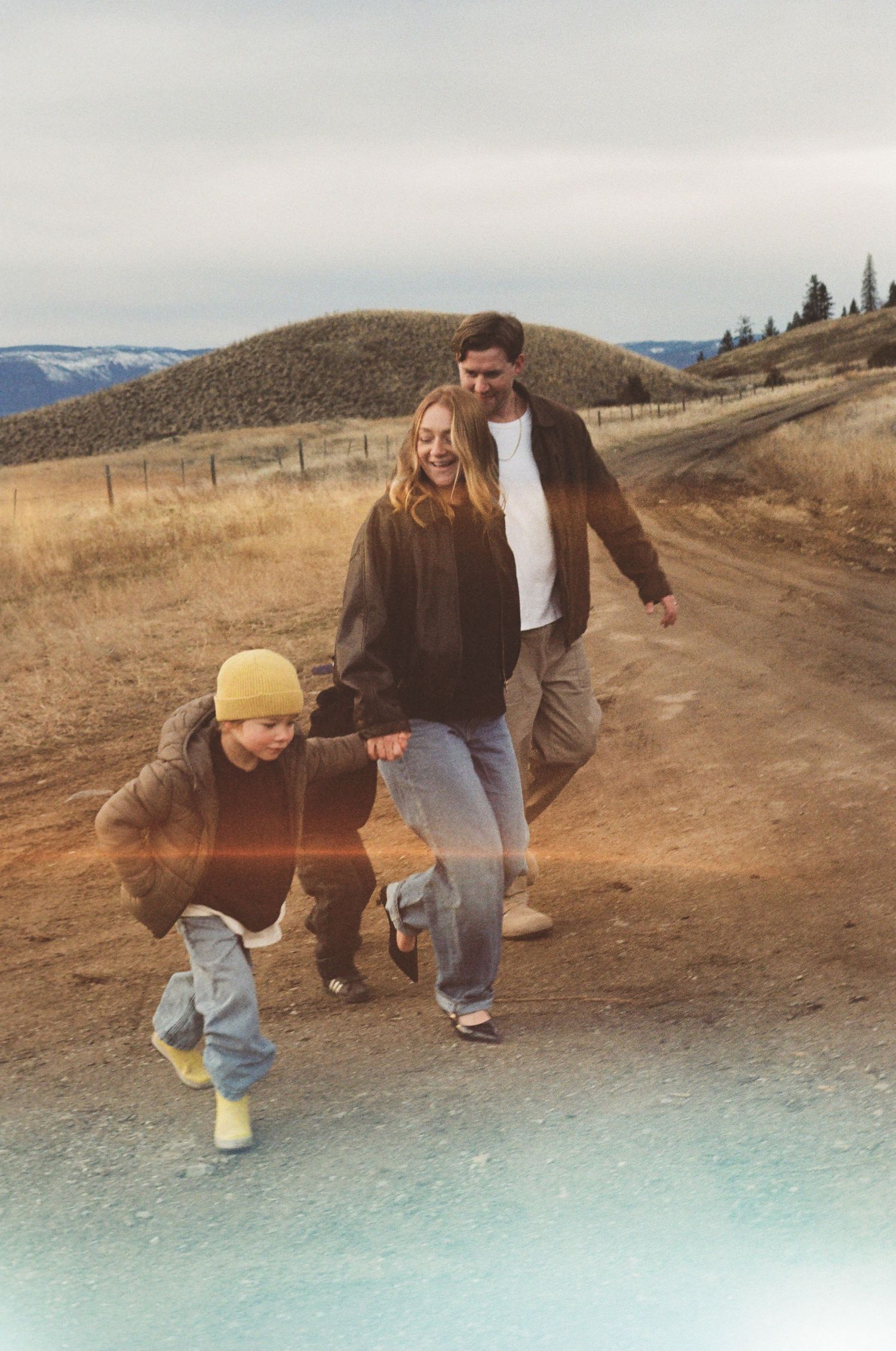 Family with young child and dog walking on rural dirt path with rolling hills in background.