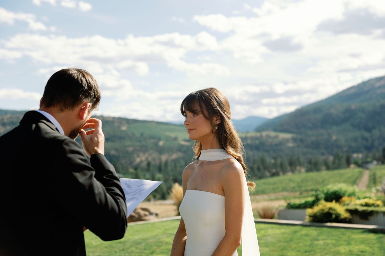 Emotional groom reads vows to smiling bride with scenic mountain valley behind.
