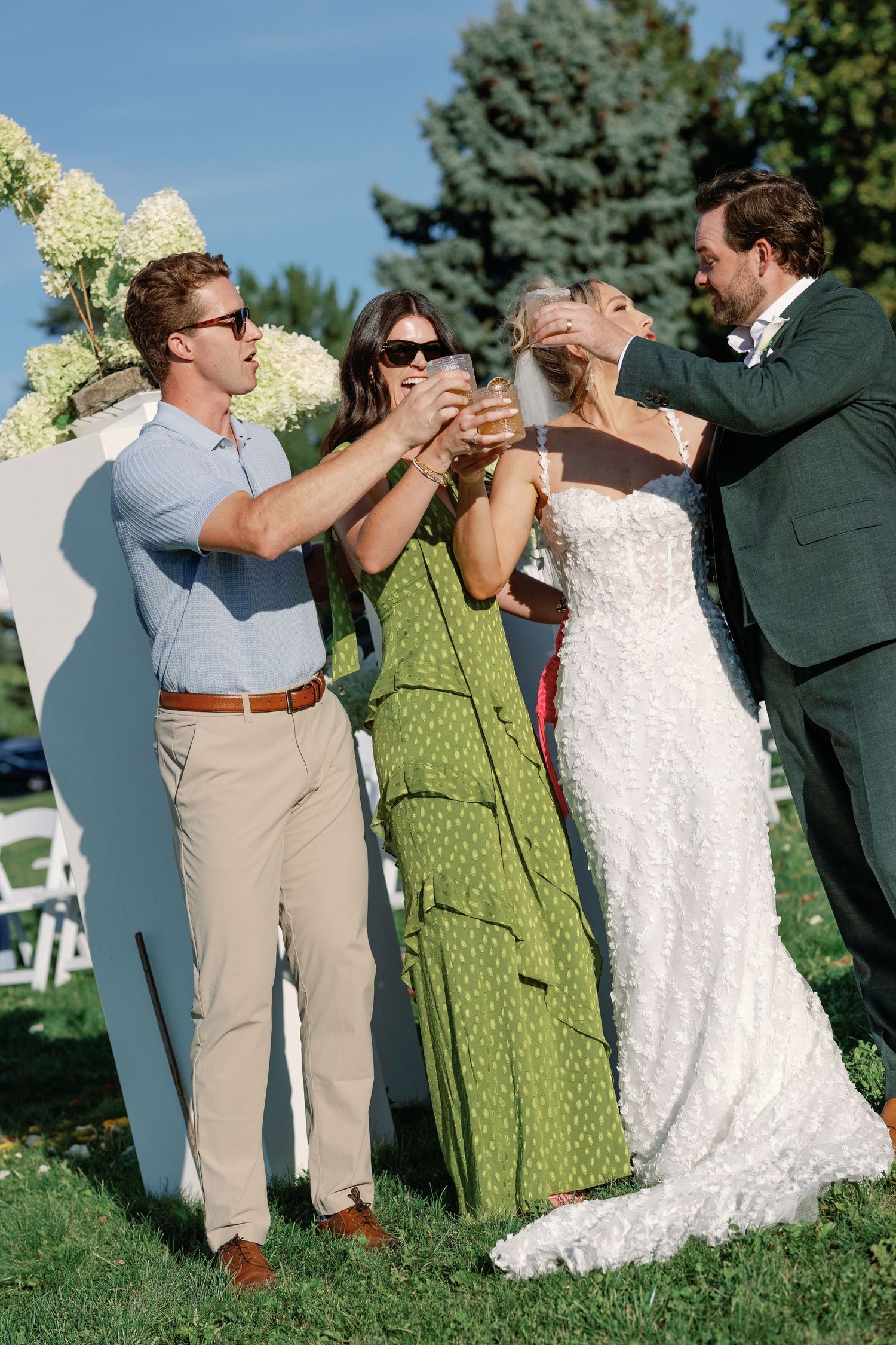 Bride and wedding guests joyfully toasting with drinks beside floral arrangements at outdoor ceremony.