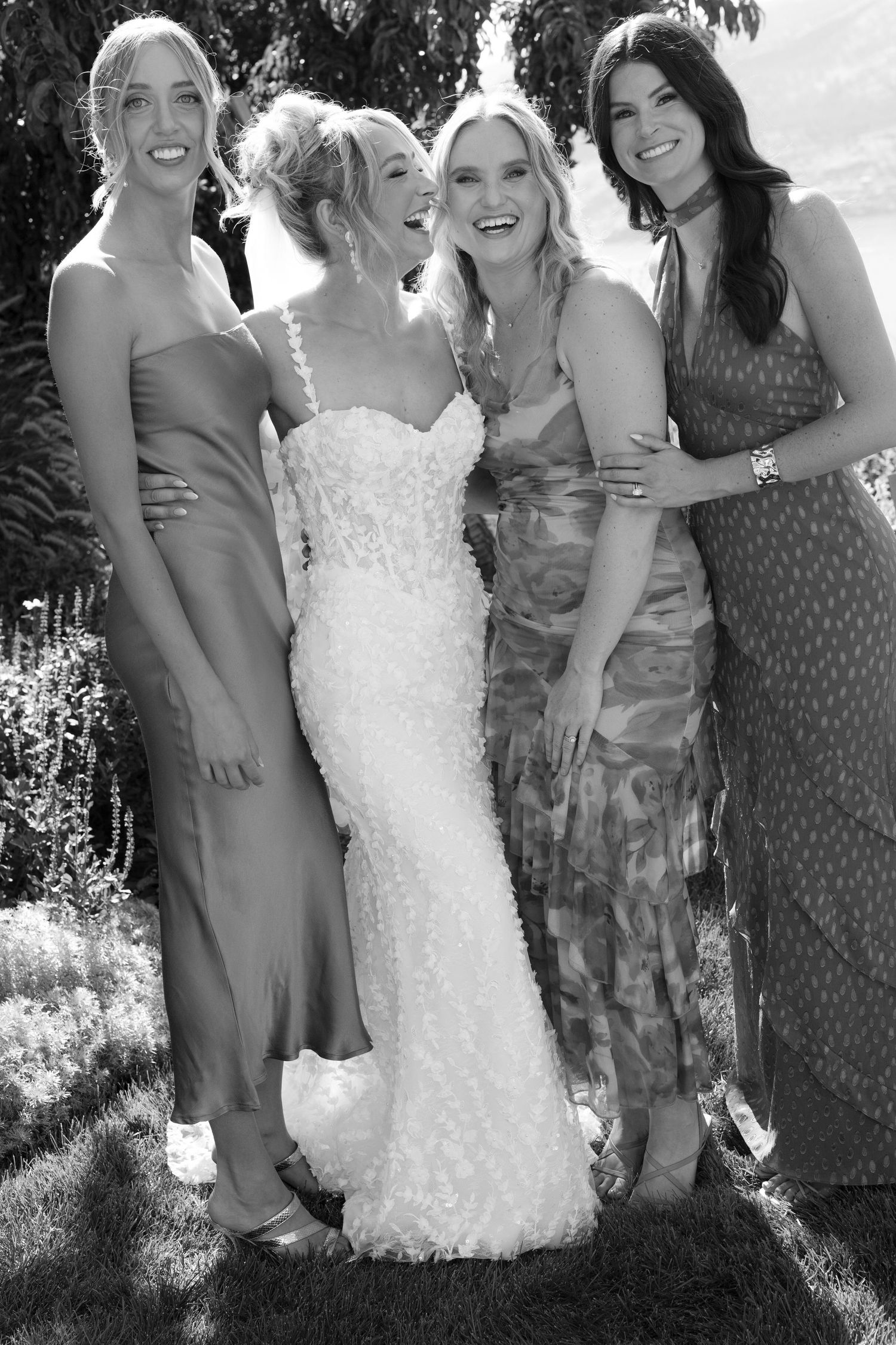Black and white photo of smiling bride in textured gown embraced by three bridesmaids in mismatched dresses.