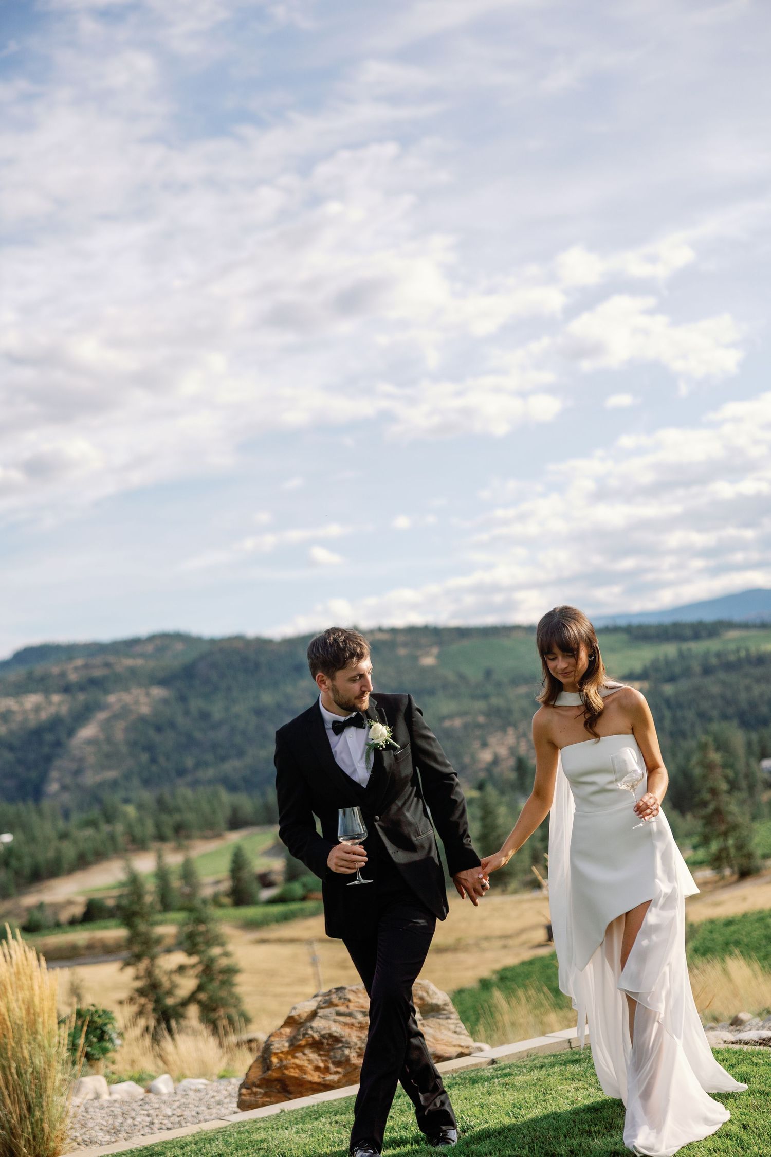 Bride in white asymmetrical gown and groom in black tuxedo walk hand in hand with panoramic mountain views.