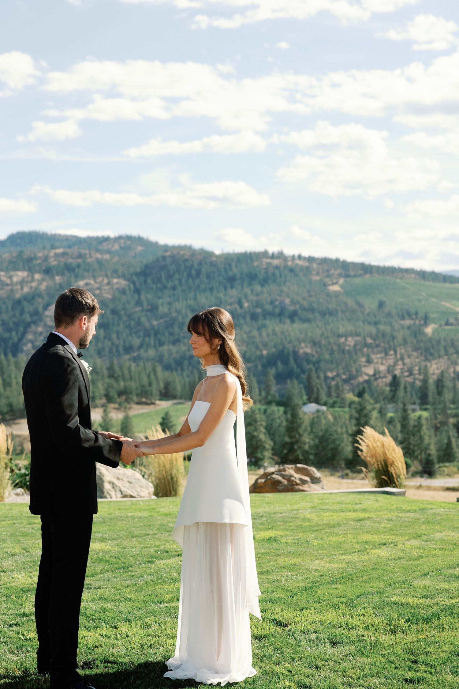 Bride in modern white gown and groom in black suit hold hands during outdoor mountain wedding ceremony.