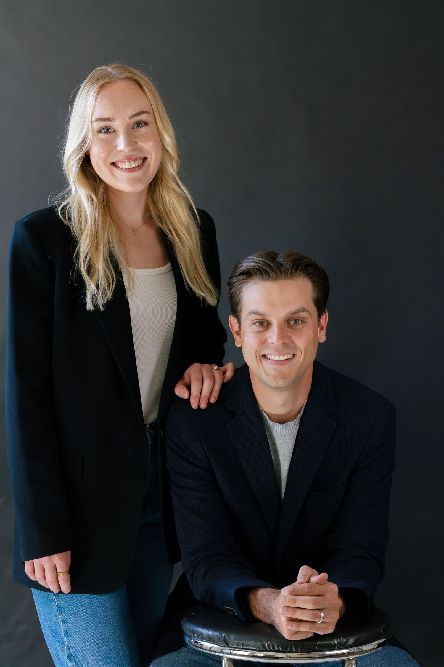 A smiling blonde woman and a man in black blazers pose together against a dark gray background.
