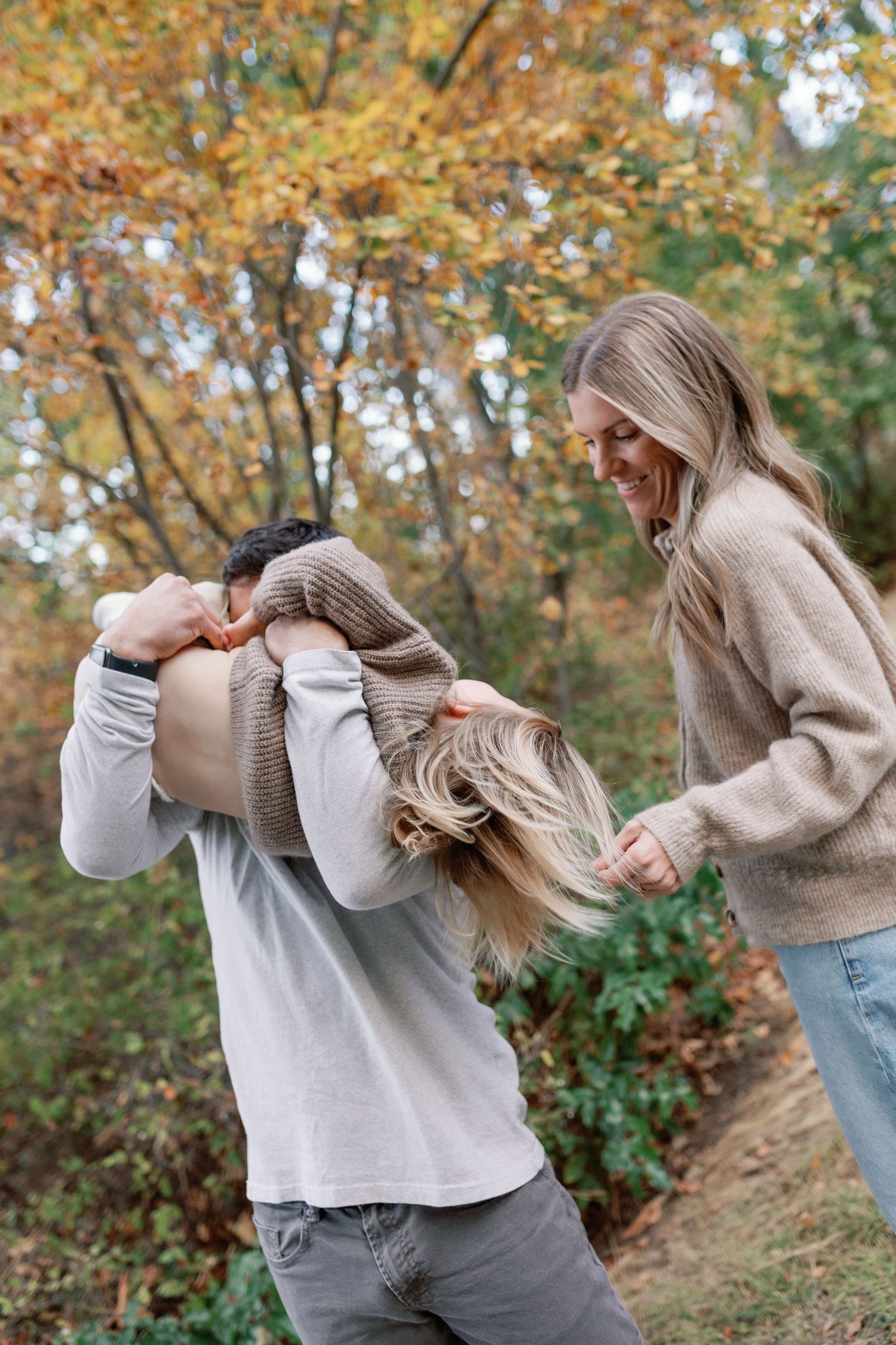Couple playfully laughing outdoors in autumn, woman smiling as man lifts her sweater over her head among colorful fall trees.