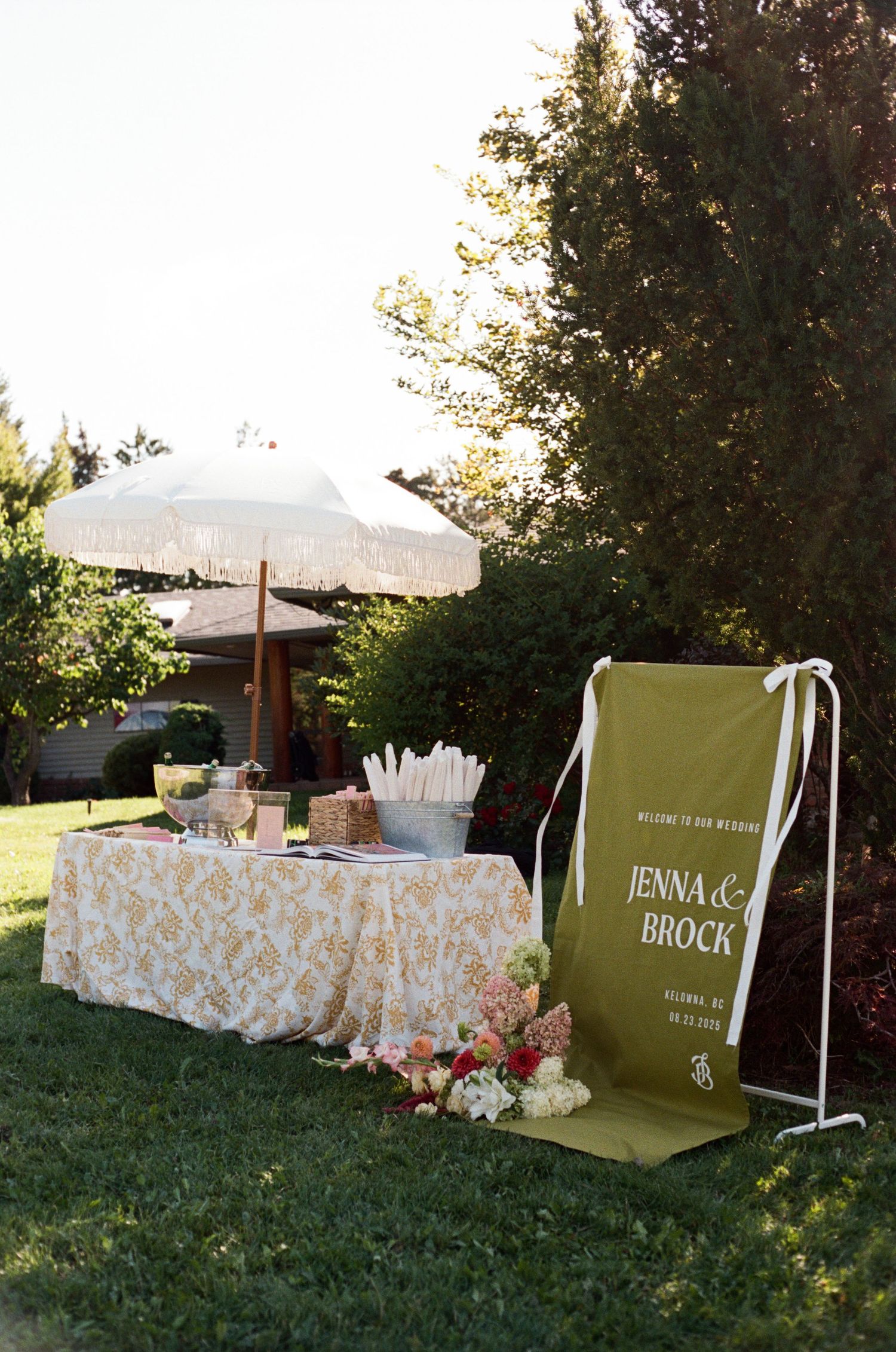 Outdoor wedding welcome sign for Jenna and Brock with floral tablecloth and umbrella in garden setting.