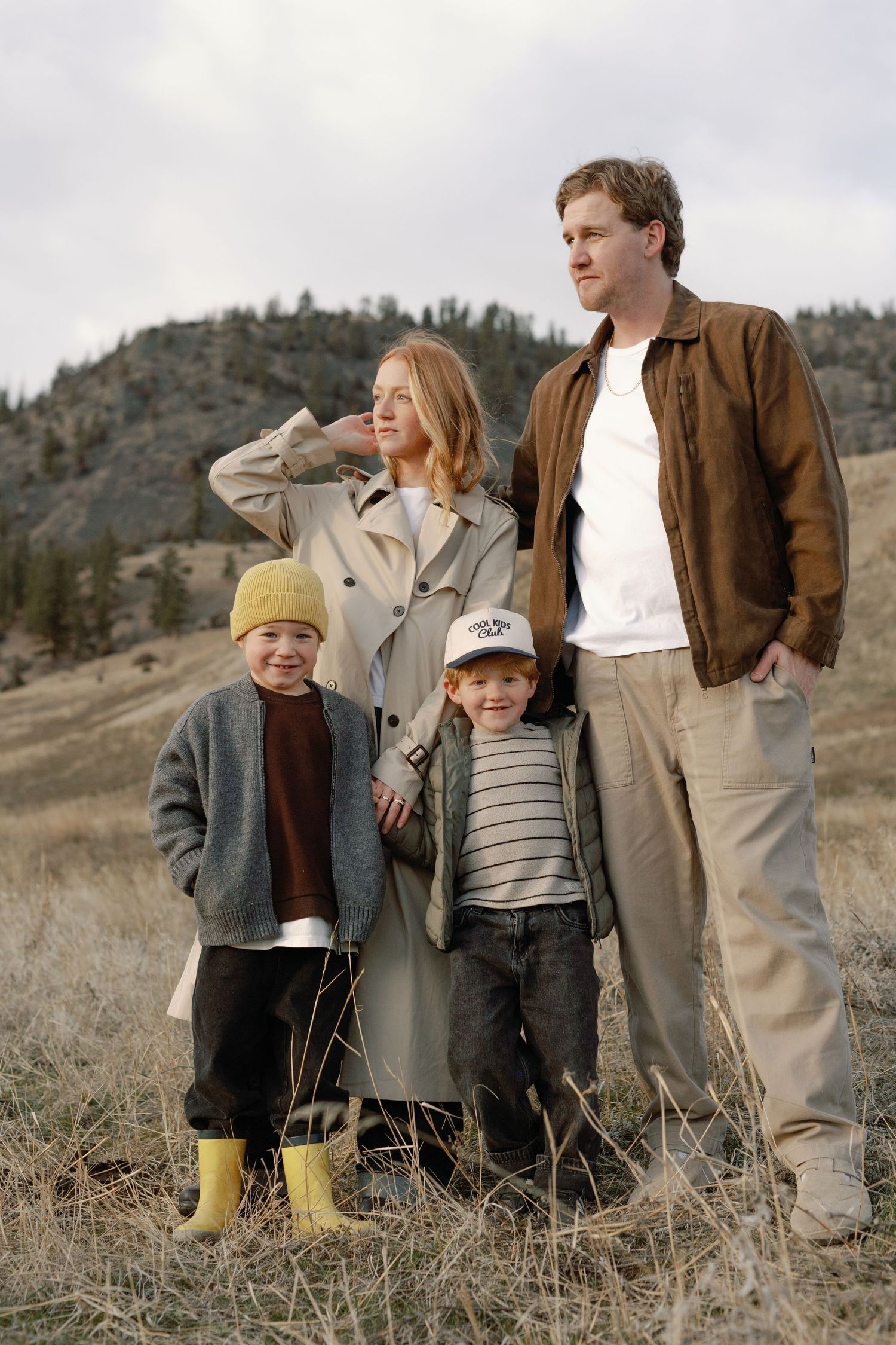 Family of four posing outdoors in a mountain meadow, wearing casual fall clothing, with hills and dry grass in background.