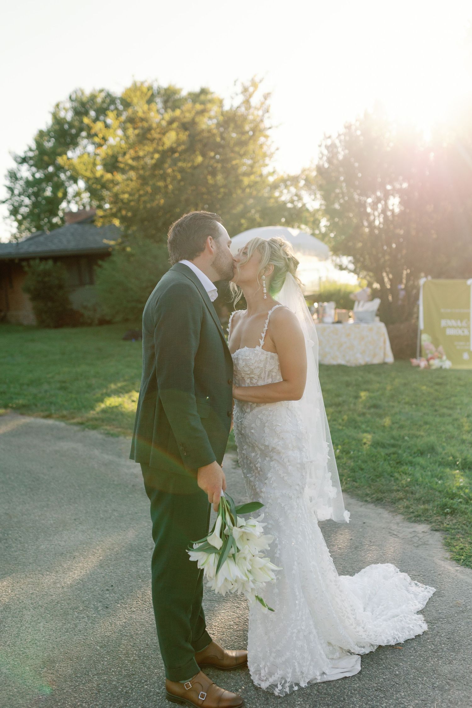 Bride and groom share a golden hour kiss on pathway holding white lilies.