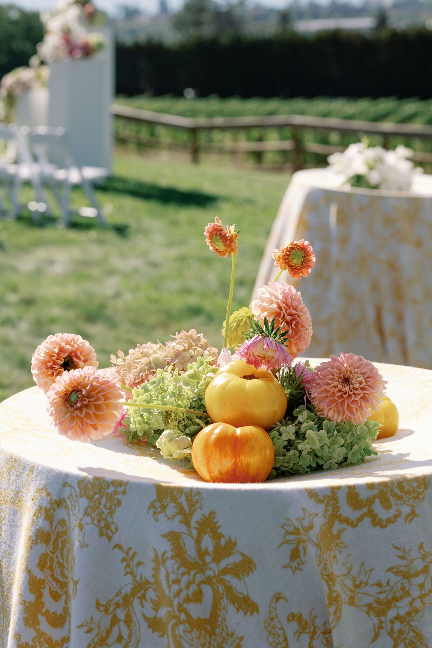 Pink dahlias and small pumpkins centerpiece on gold floral tablecloth at outdoor wedding reception.