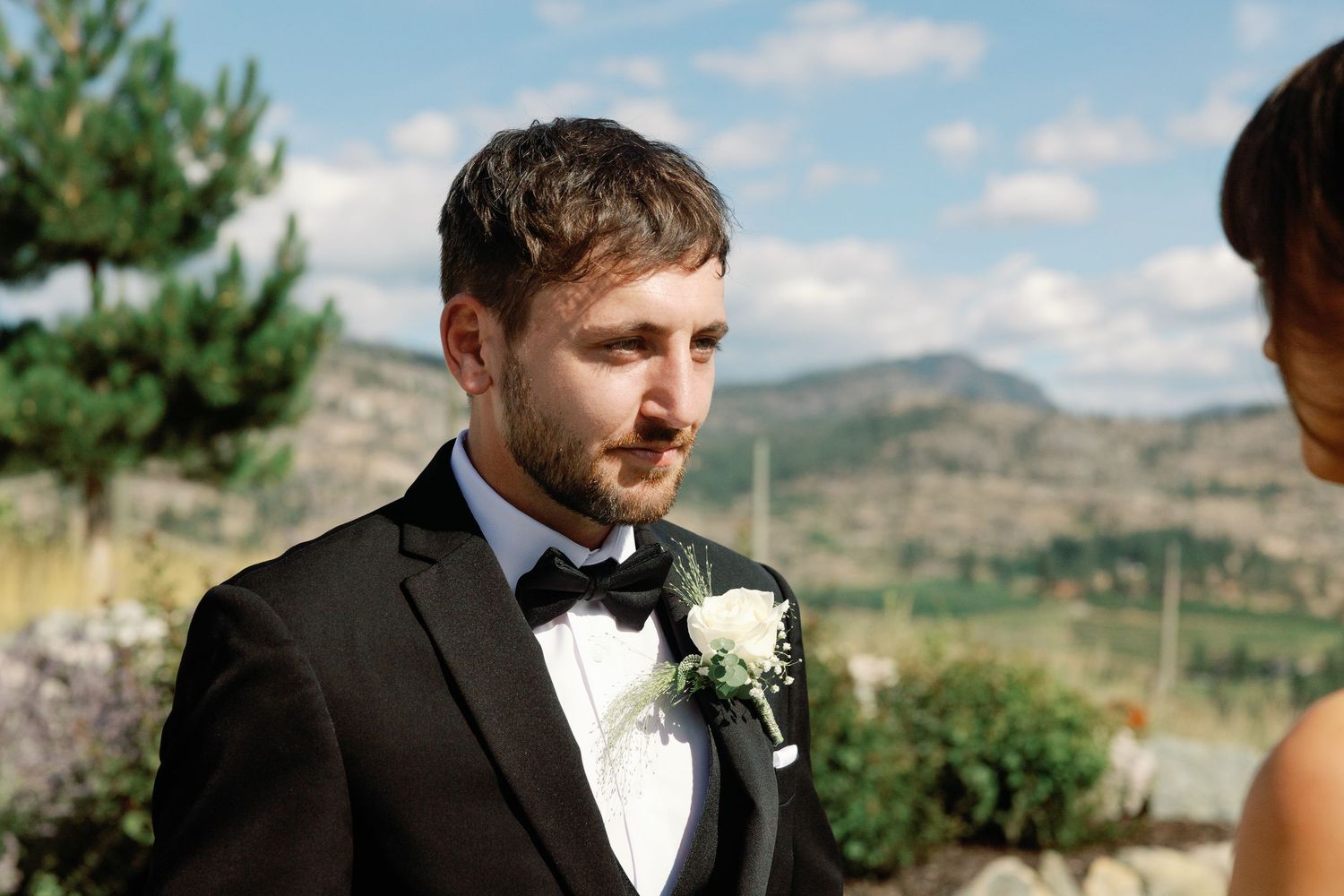 Emotional groom in black tuxedo with white boutonniere listens to vows during outdoor ceremony.