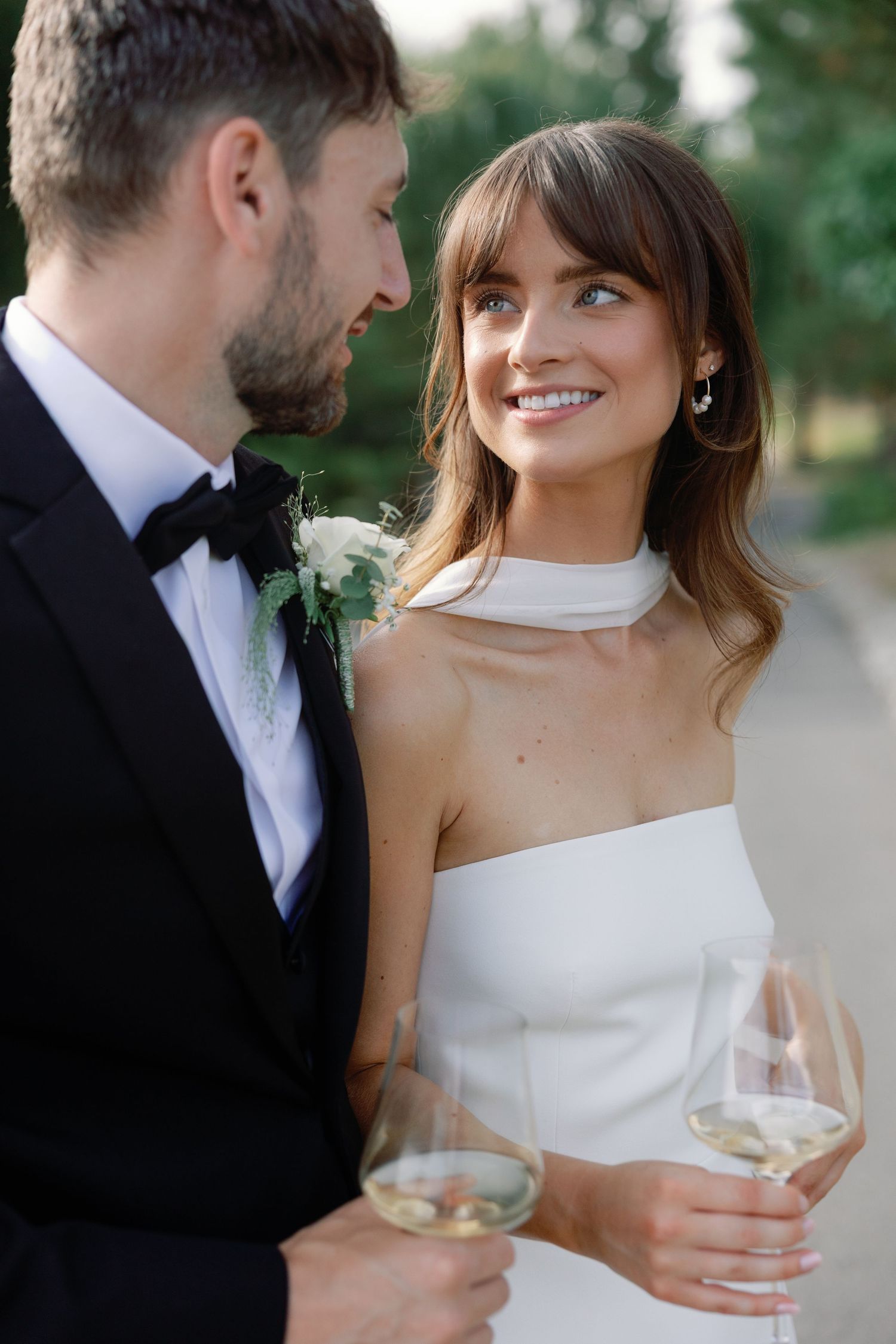 Smiling bride with bangs in white halter gown gazes at groom while holding champagne glasses outdoors.