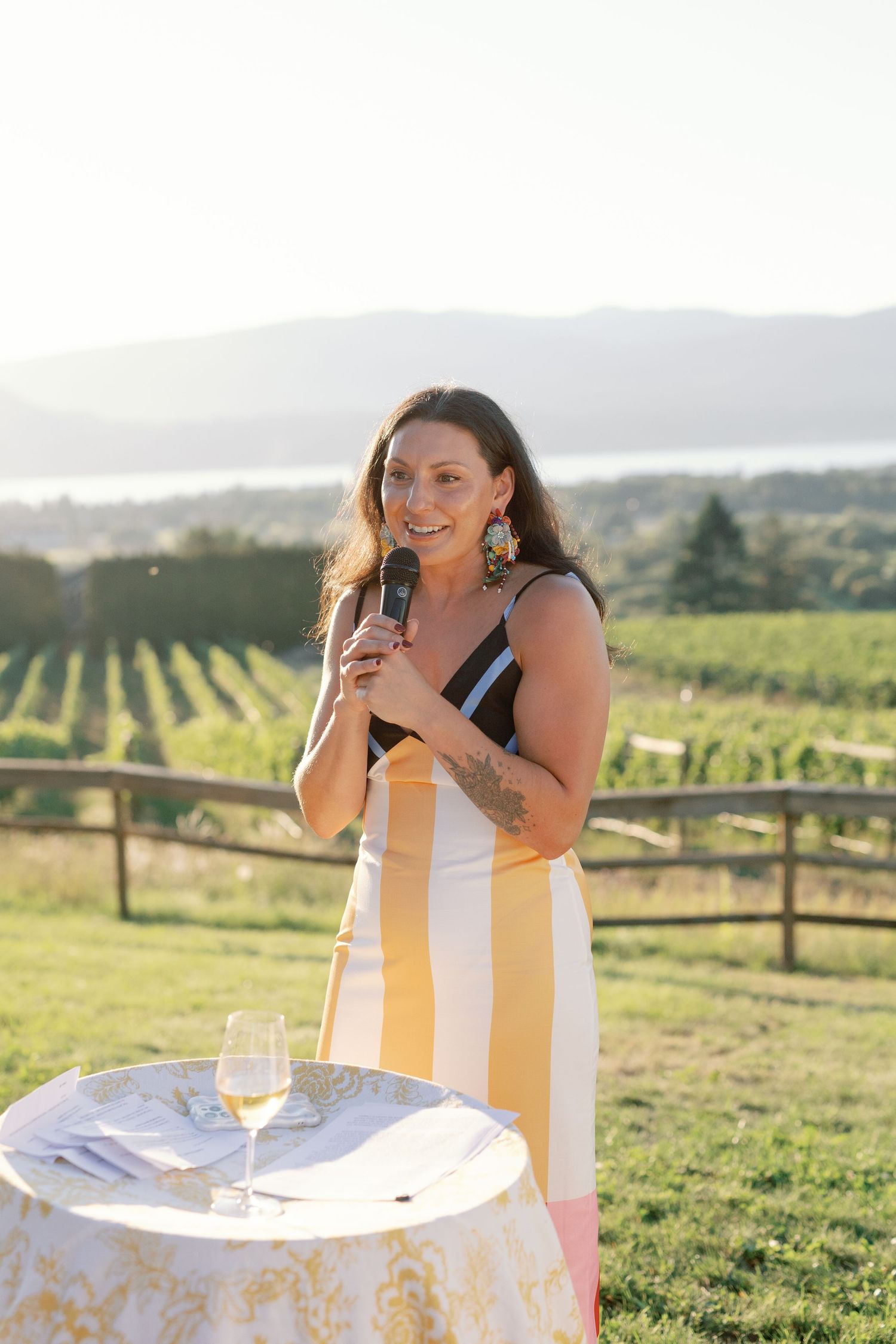 Woman in yellow striped dress gives speech at vineyard wedding with scenic mountain backdrop.