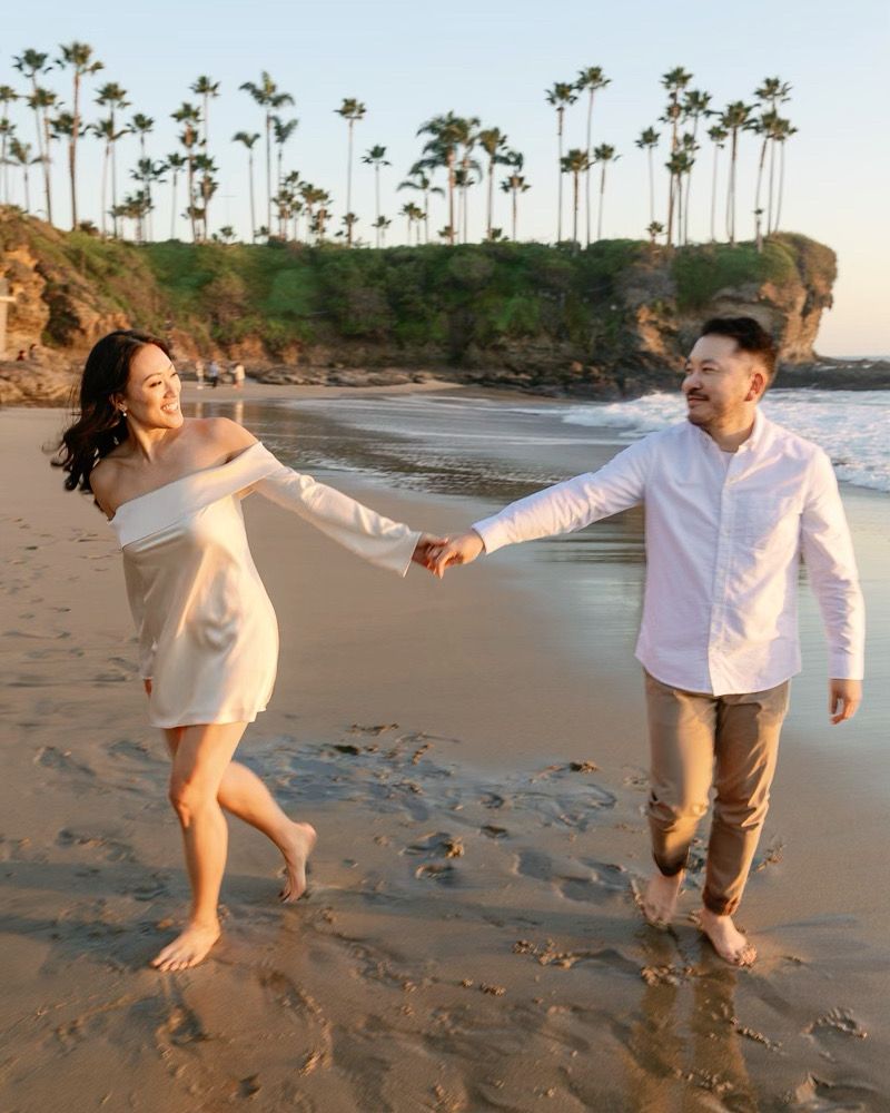 A joyful couple holds hands and walks along a scenic beach with palm trees and rocky cliffs at sunset.