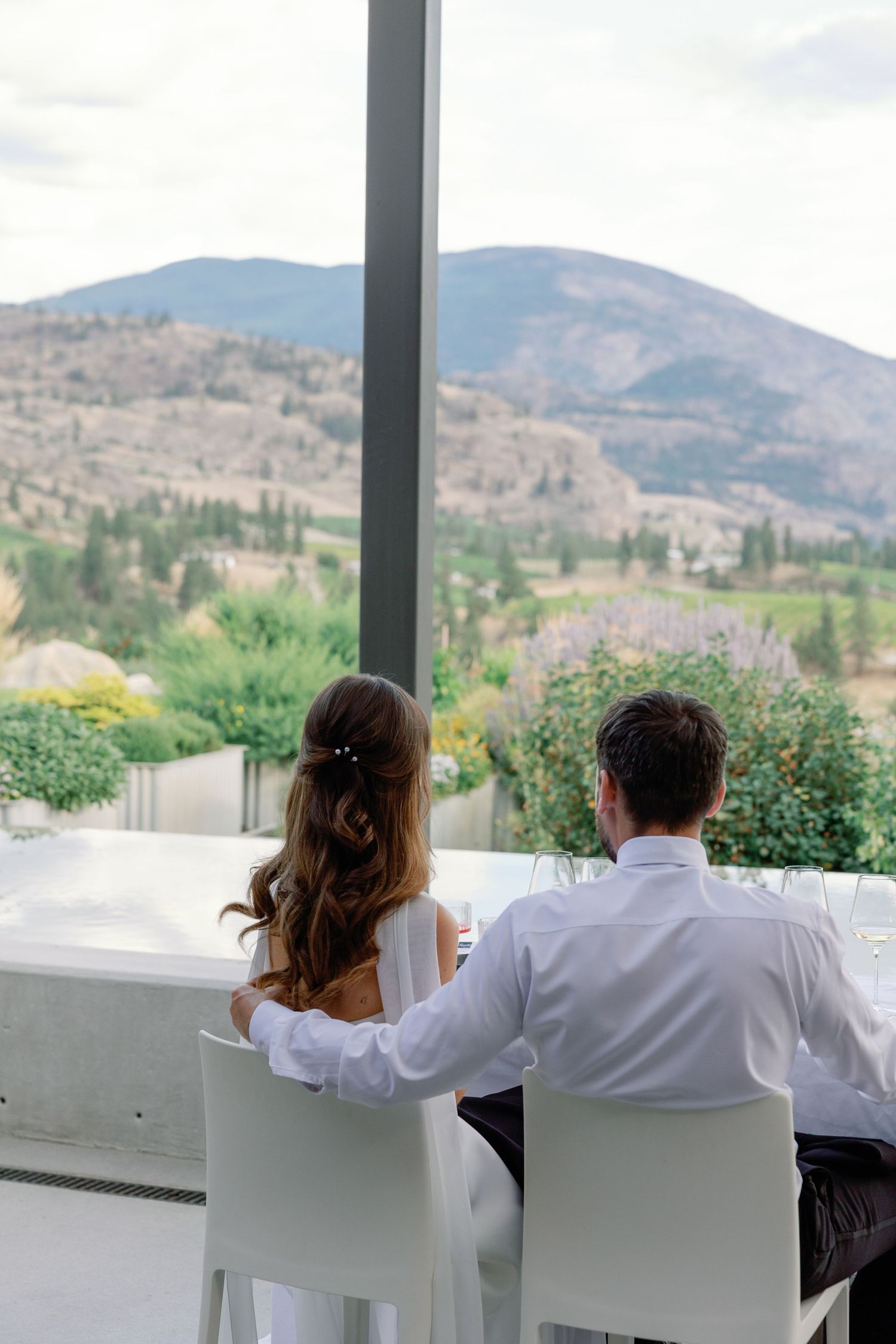 Bride and groom seated at a white dining table admiring a stunning mountain landscape through floor-to-ceiling windows.