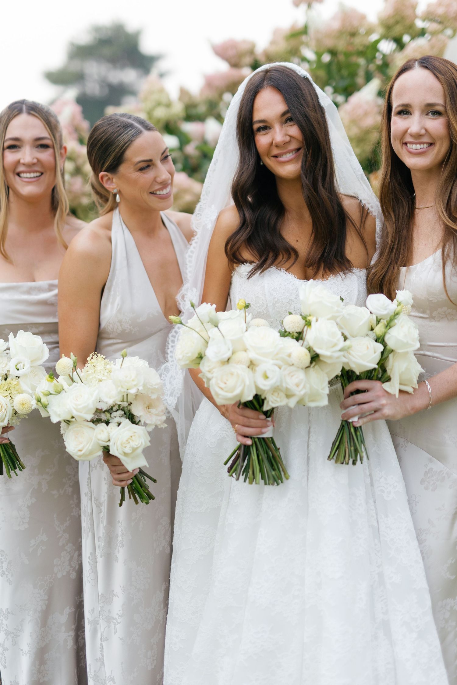 Bride in white gown and veil holds bouquet with three bridesmaids in silver dresses at outdoor wedding.