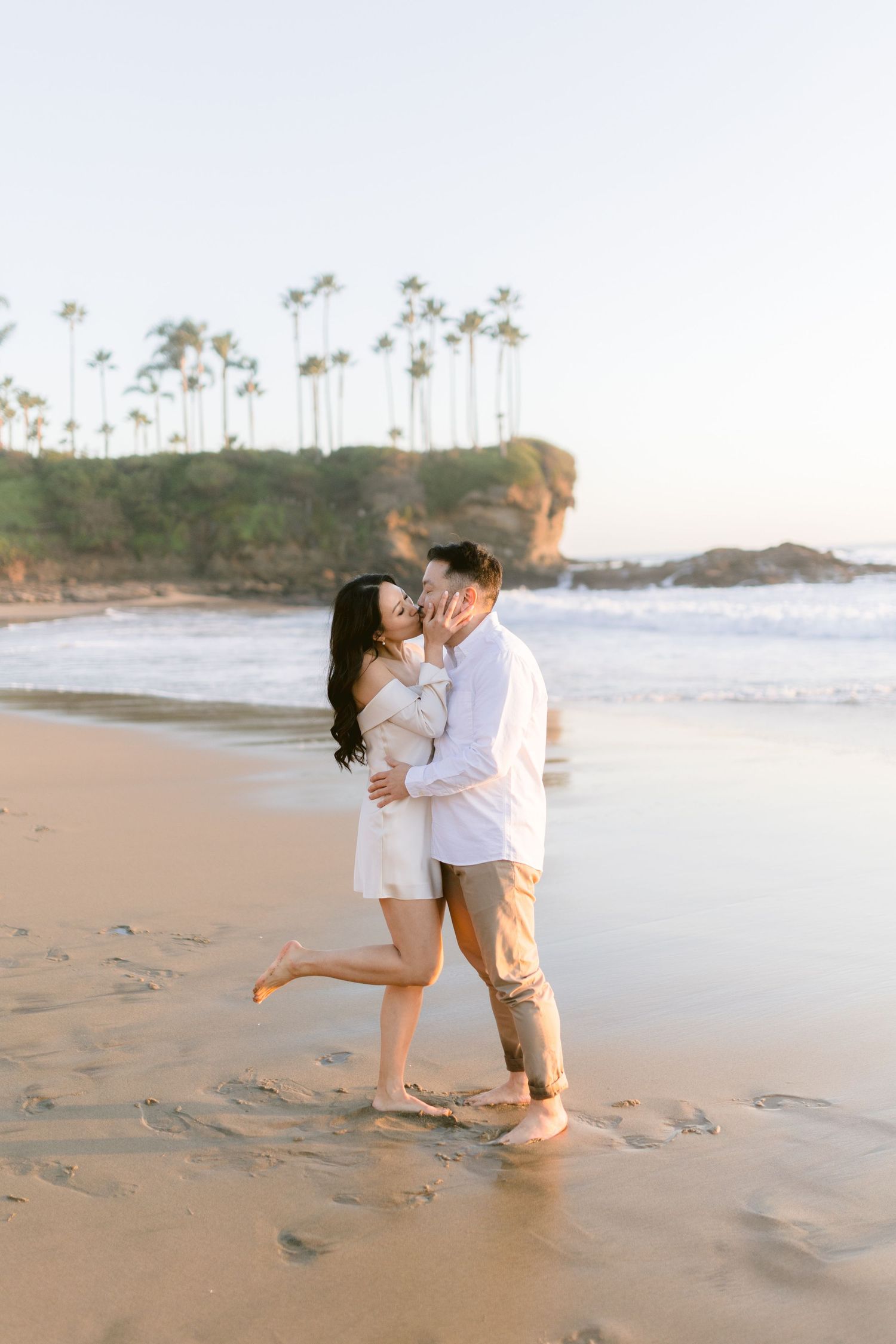 A couple shares a romantic kiss on a sandy beach at sunset with palm trees and ocean waves behind them.