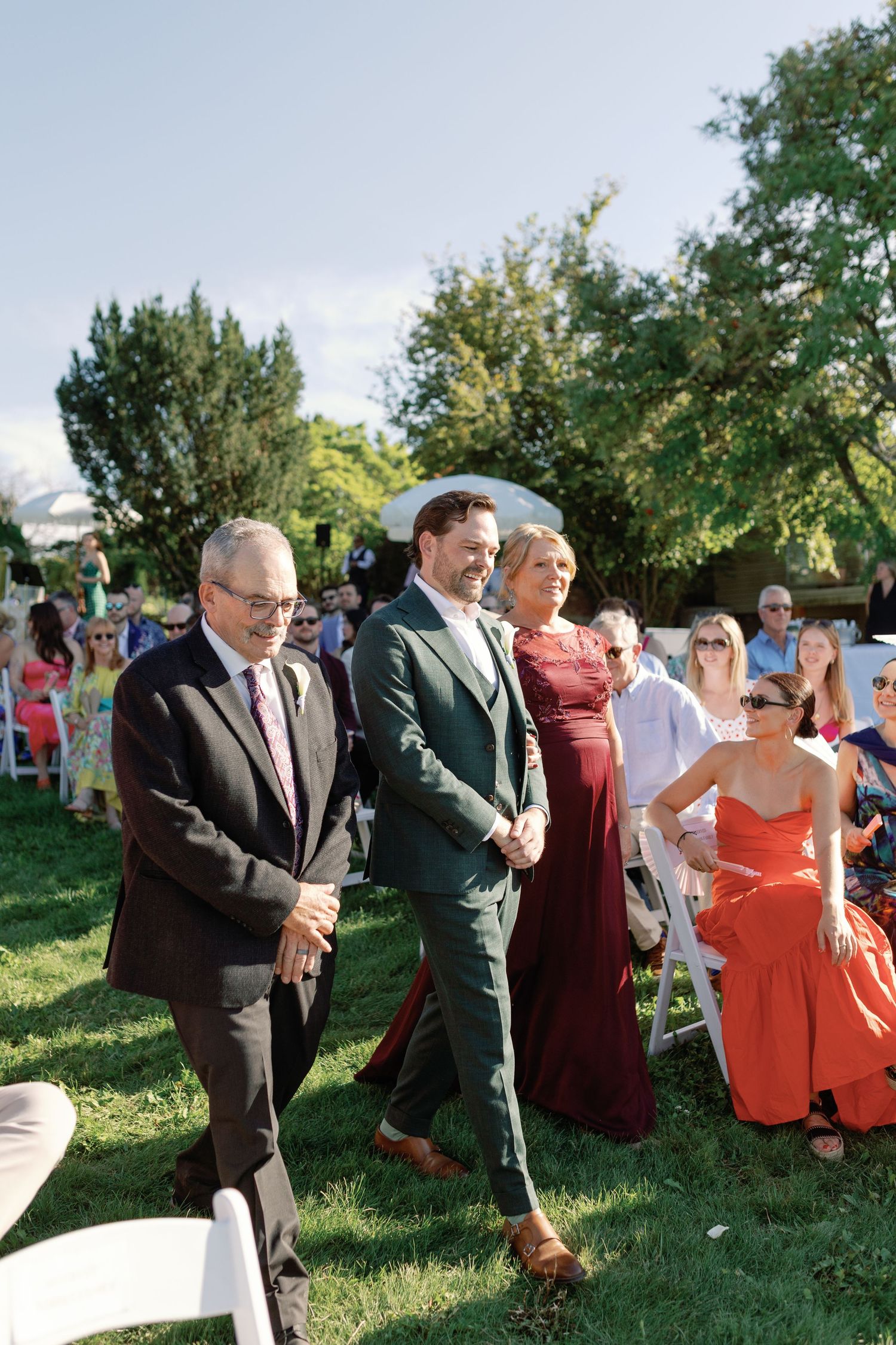 Groom in green suit walking down the aisle with parents at an outdoor wedding ceremony.